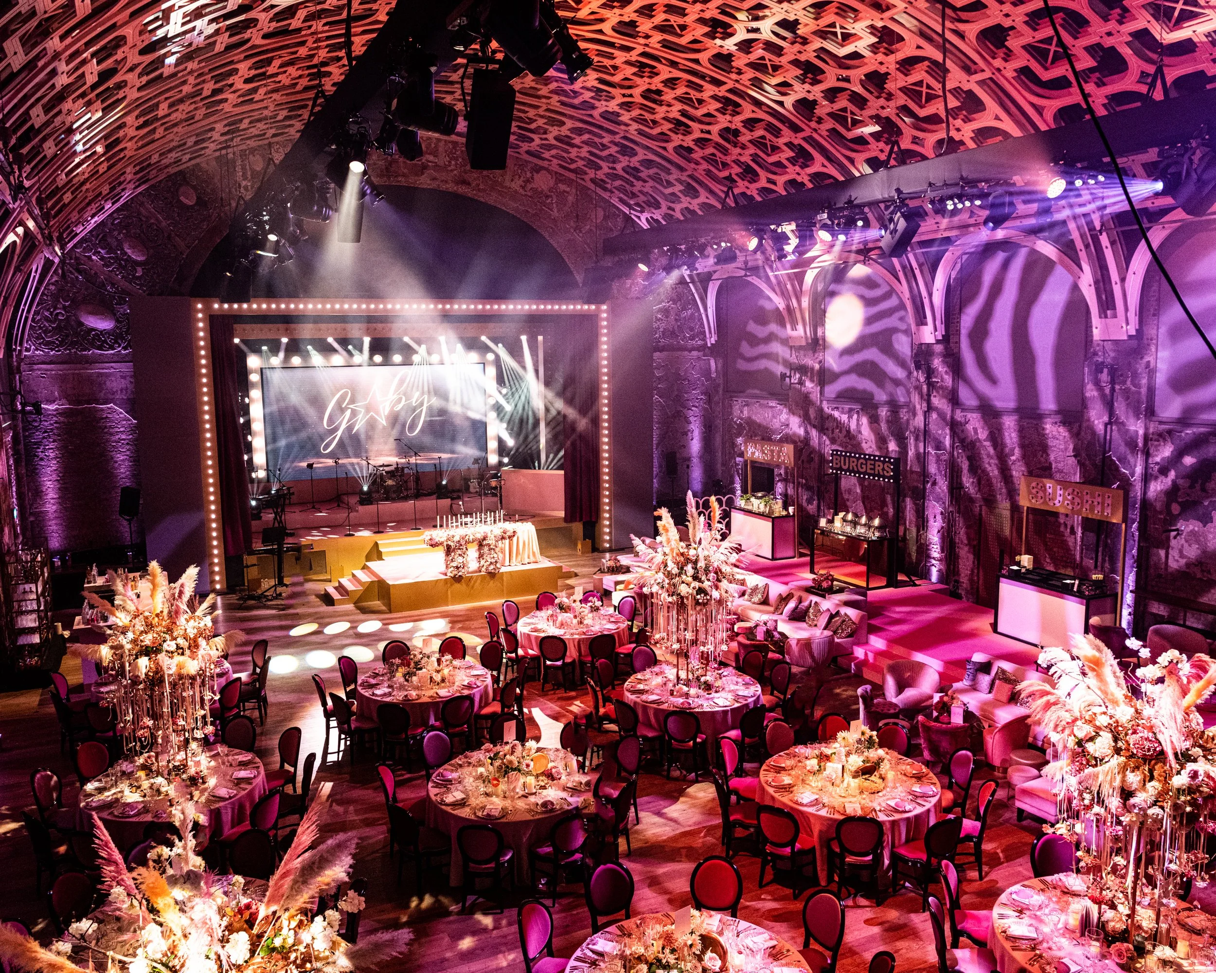 An elegant event space with round tables decorated with floral centerpieces and candles, a decorated stage with lighting, and pink and purple ambient lighting highlighting ornate architectural features. BAT MITZVAH AT BATTERSEA ARTS CENTRE