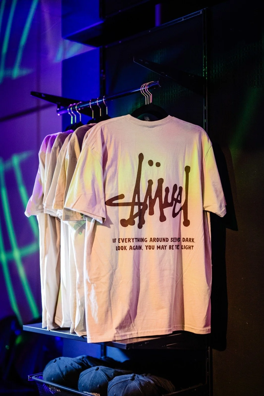 Pink T-shirt with black graffiti-style text and quote hanging among other T-shirts on a clothing rack, in a store with colorful neon lighting.