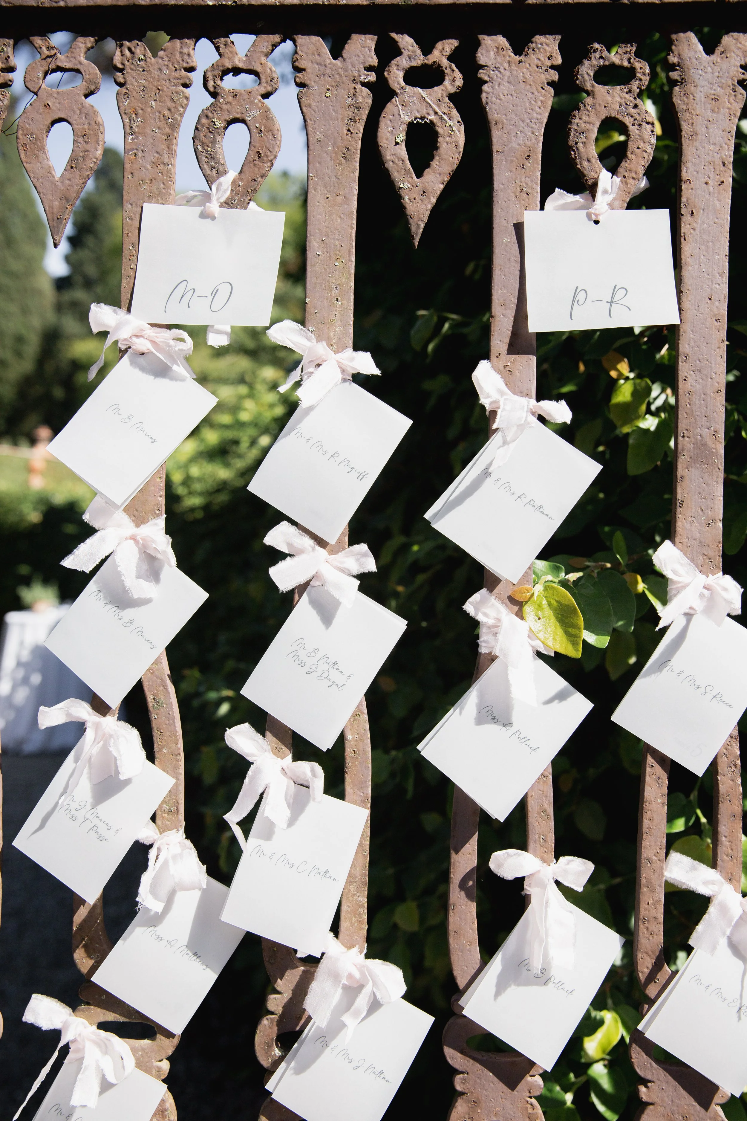 A decorative wrought iron fence with white cards tied to it with white cloth ribbons. The cards have handwritten names for the weddings  seating chart. There is greenery in the background.