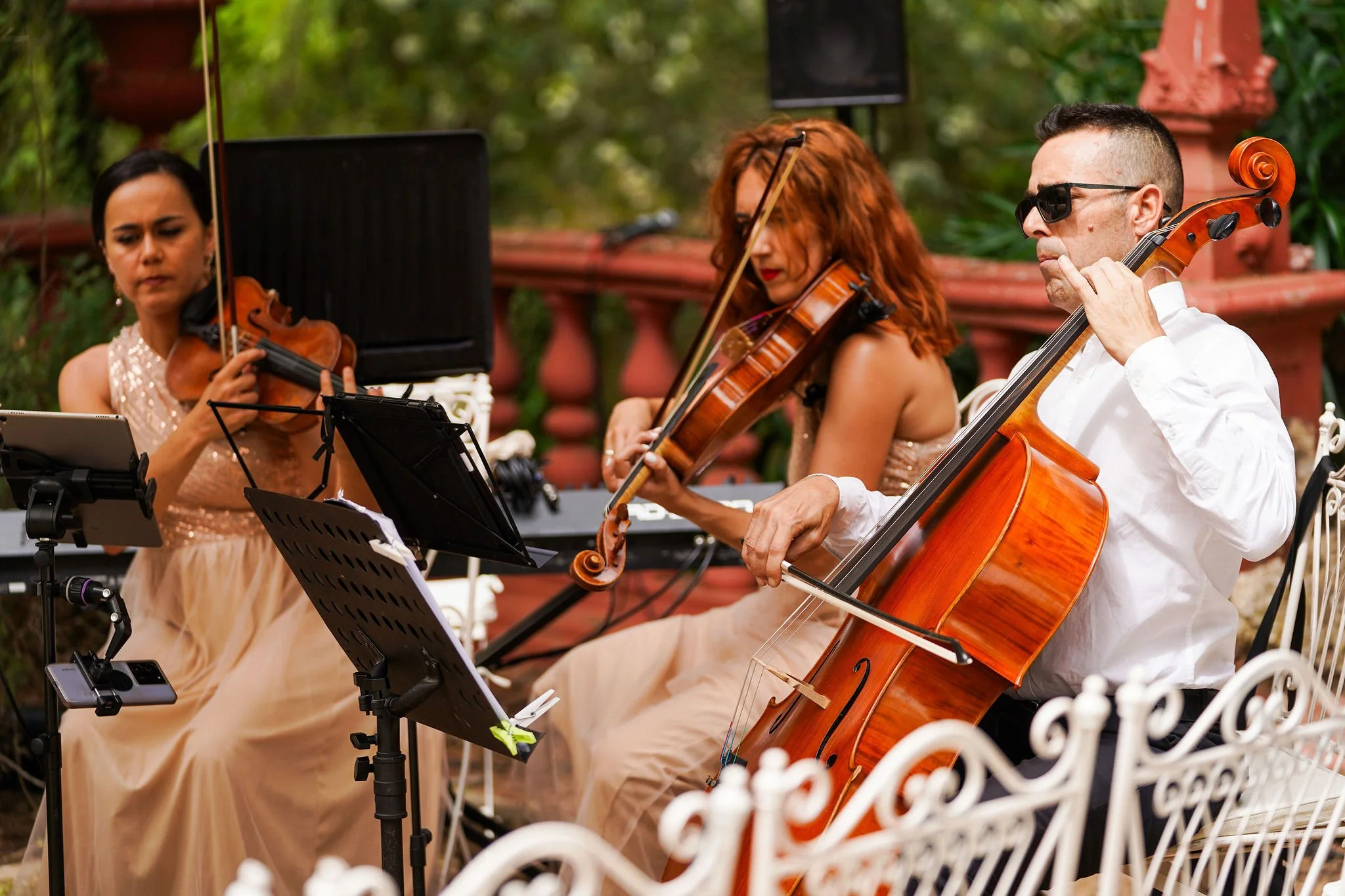 Three musicians playing string instruments outdoors, two women on the left and a man on the right, with a white decorative fence in the foreground and green foliage in the background at Anantara Villa Padierna Resort in Marbella