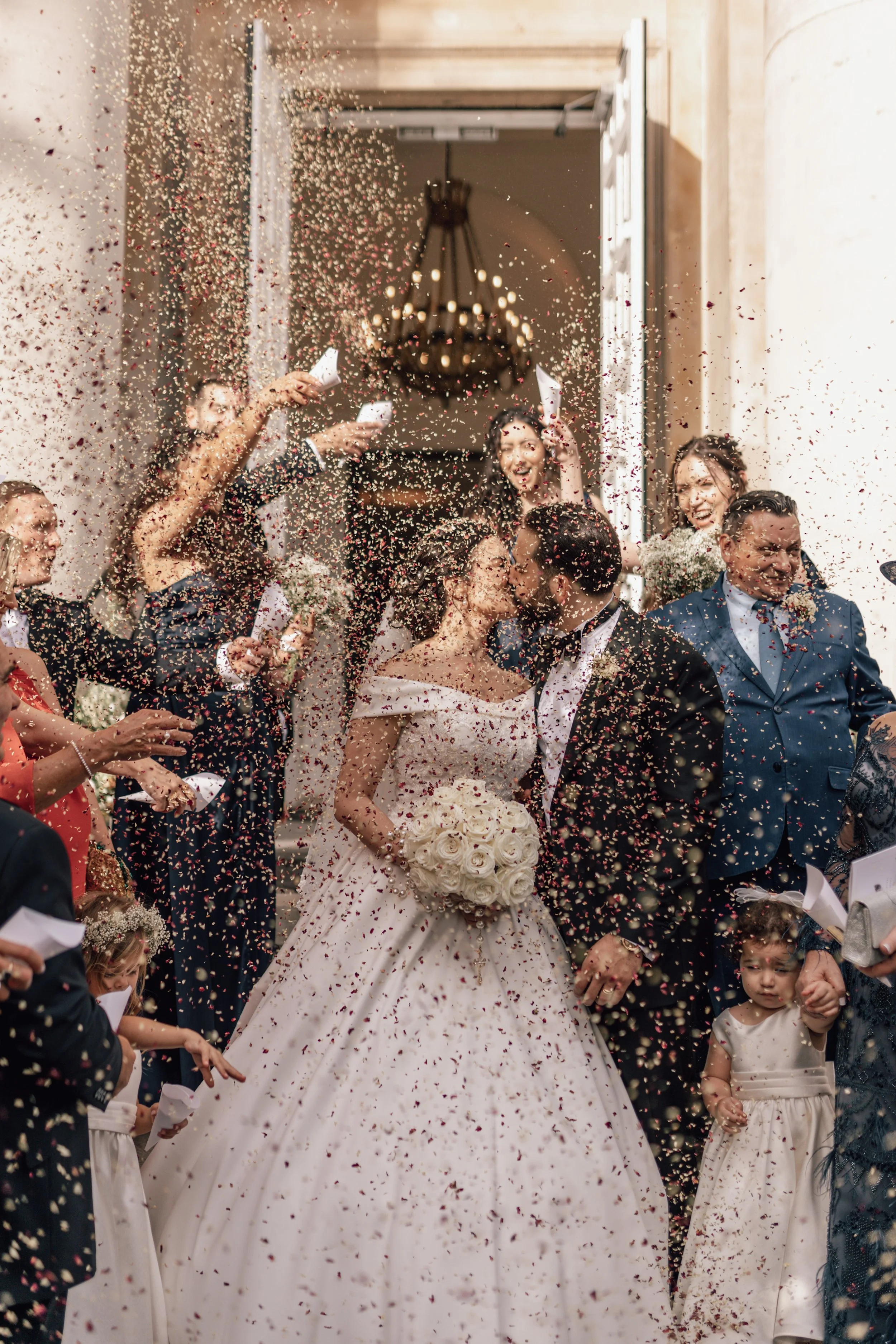 A bride and groom kiss as guests throw confetti during their wedding celebration outside a building with an open door and chandelier.