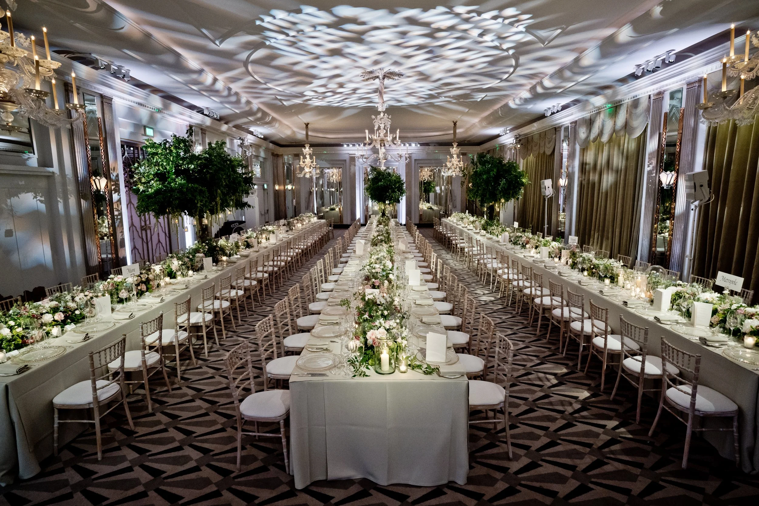 Elegant banquet hall decorated for a wedding reception, with long tables adorned with white tablecloths, floral centerpieces, candles, and surrounded by white chairs. Large green trees and ornate chandeliers are suspended from the ceiling, with intri