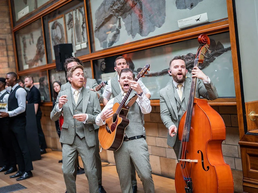 A group of men playing musical instruments at an event, with one man singing and playing the guitar, another on the double bass, and others in the background. The Natural History Museum wall with display cases of fossils or rocks.