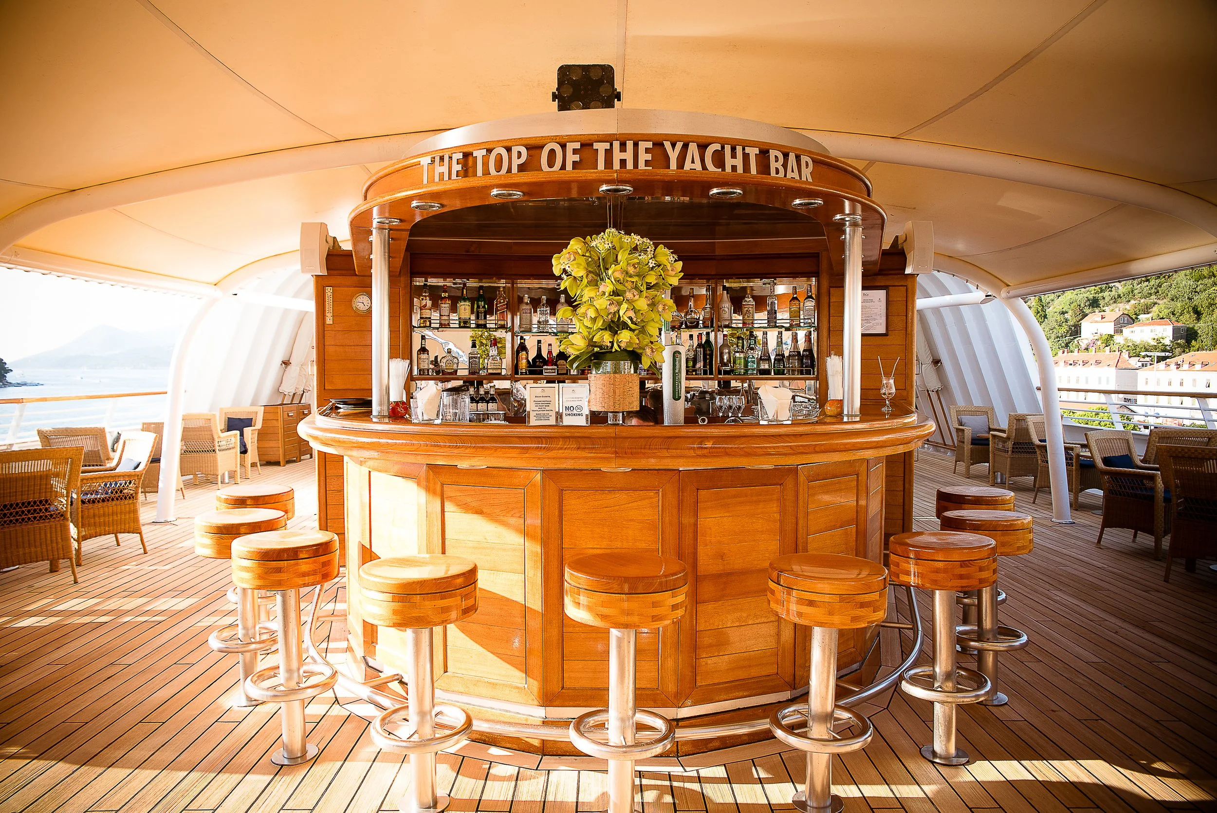 A bar located on the top deck of a yacht, with a curved wooden counter, bar stools, and a large flower arrangement on the counter. There are seating areas with chairs and tables around, and a scenic view of water and houses in the background.
