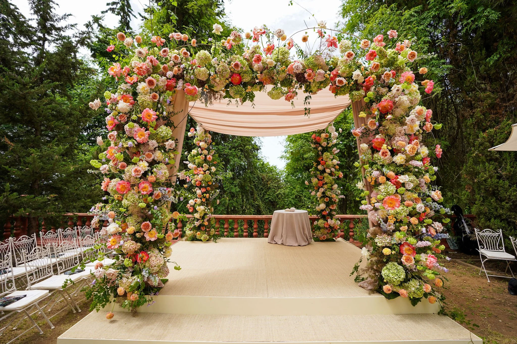 A floral wedding chuppah decorated with pink, peach, and white flowers, draped with pink fabric, set outdoors on a beige carpeted platform surrounded by trees and white chairs at Anantara Villa Padierna Resort in Marbella