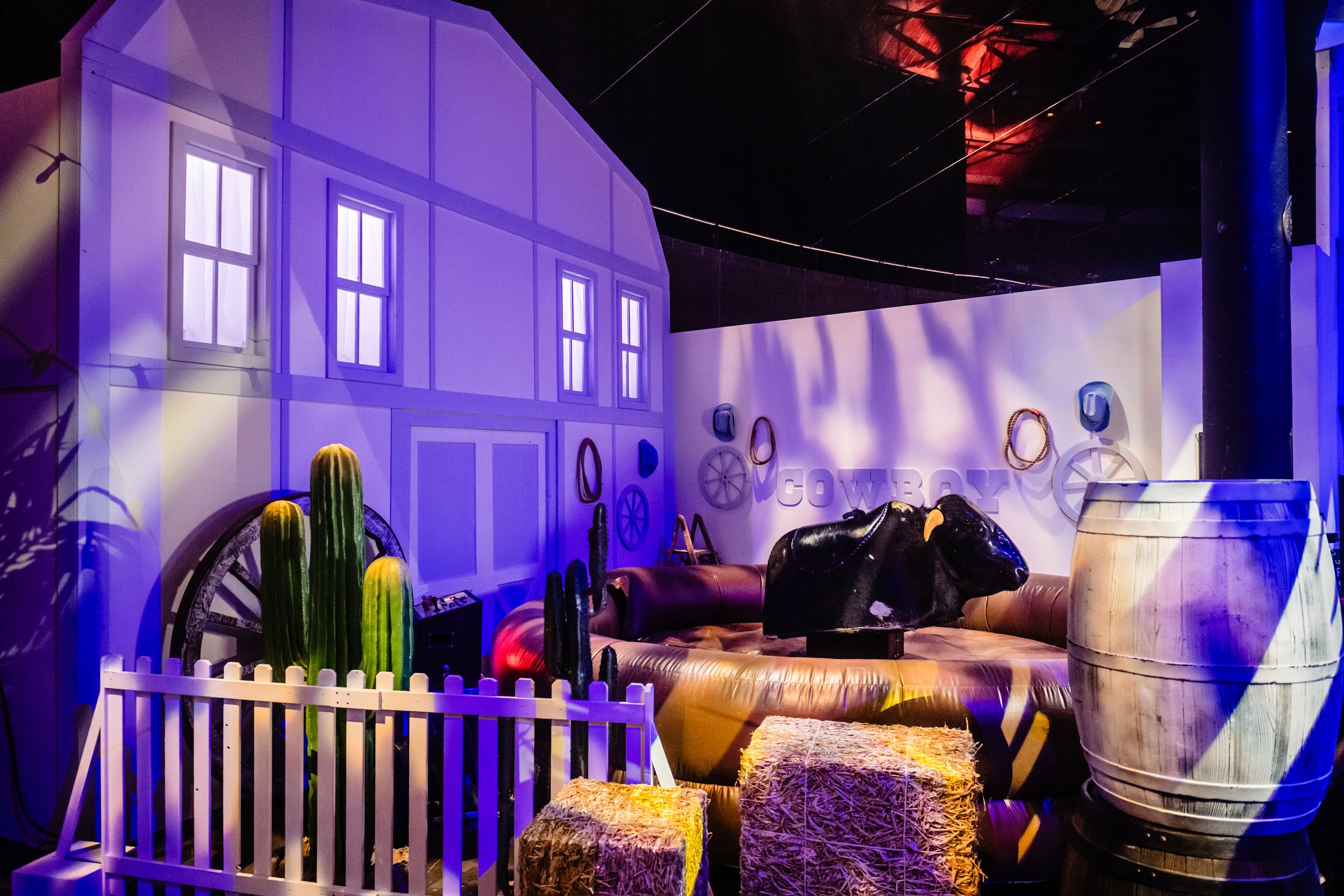 A Bar Mitzvah with a white fence, cacti, hay bales, and a life-sized black cow statue. There are building facades with windows and decorations like wagon wheels and lettered signs. 
