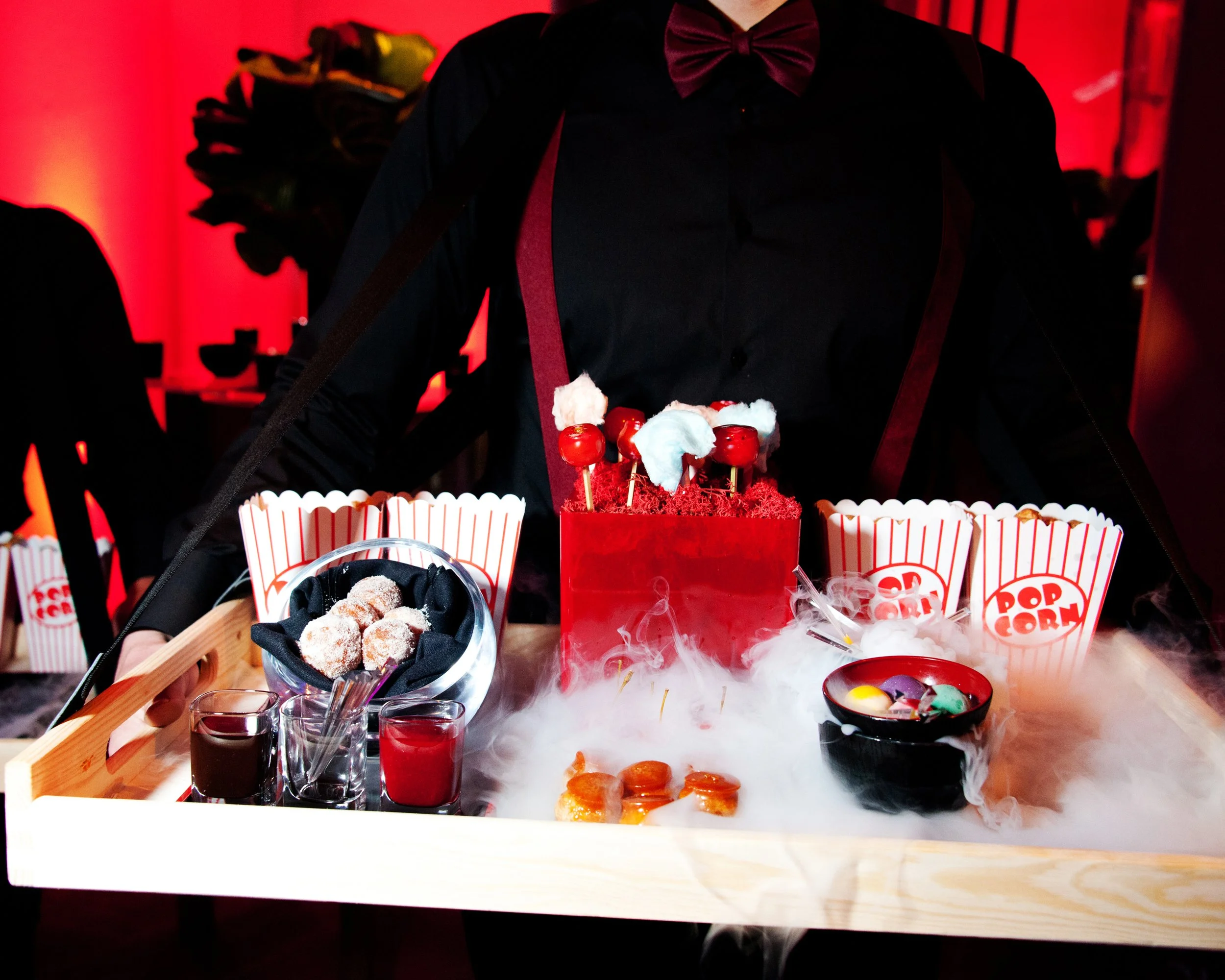 A person wearing a black shirt, red bow tie, and suspenders standing behind a table with Halloween-themed treats, including red and green candies, cupcakes, and small jars of red liquid, with a red background and Halloween decorations.