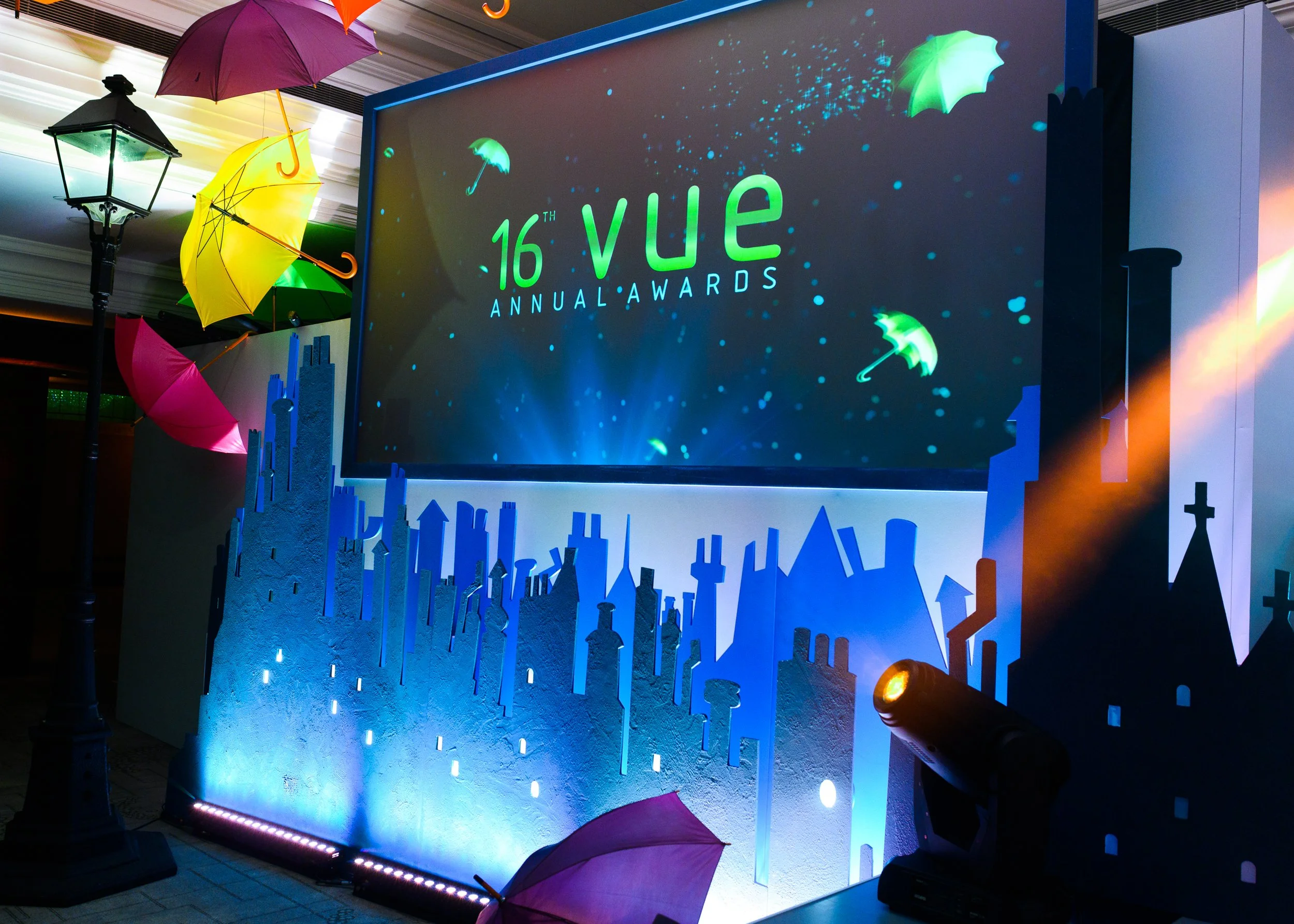 Large digital display at the 16th VUE Annual Awards, illuminated with blue and green lights. Several colorful umbrellas and a vintage-style street lamp decorate the scene. The display features the event's name and logo.