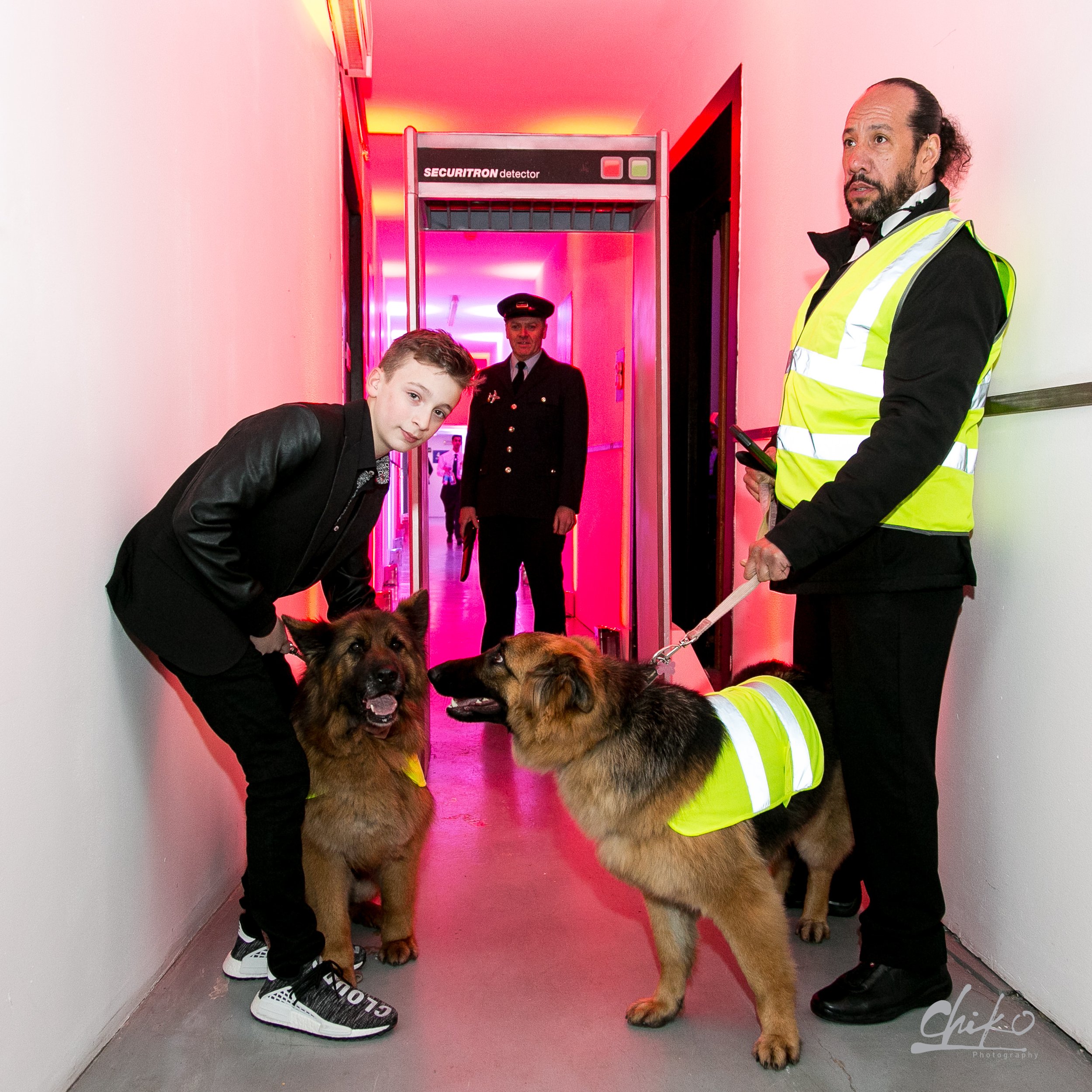 A young man with a dog and a woman with a dog in a hallway with pink lighting. Two uniformed officers are in the background near an entry gate labeled 'SECURITRON detector'.