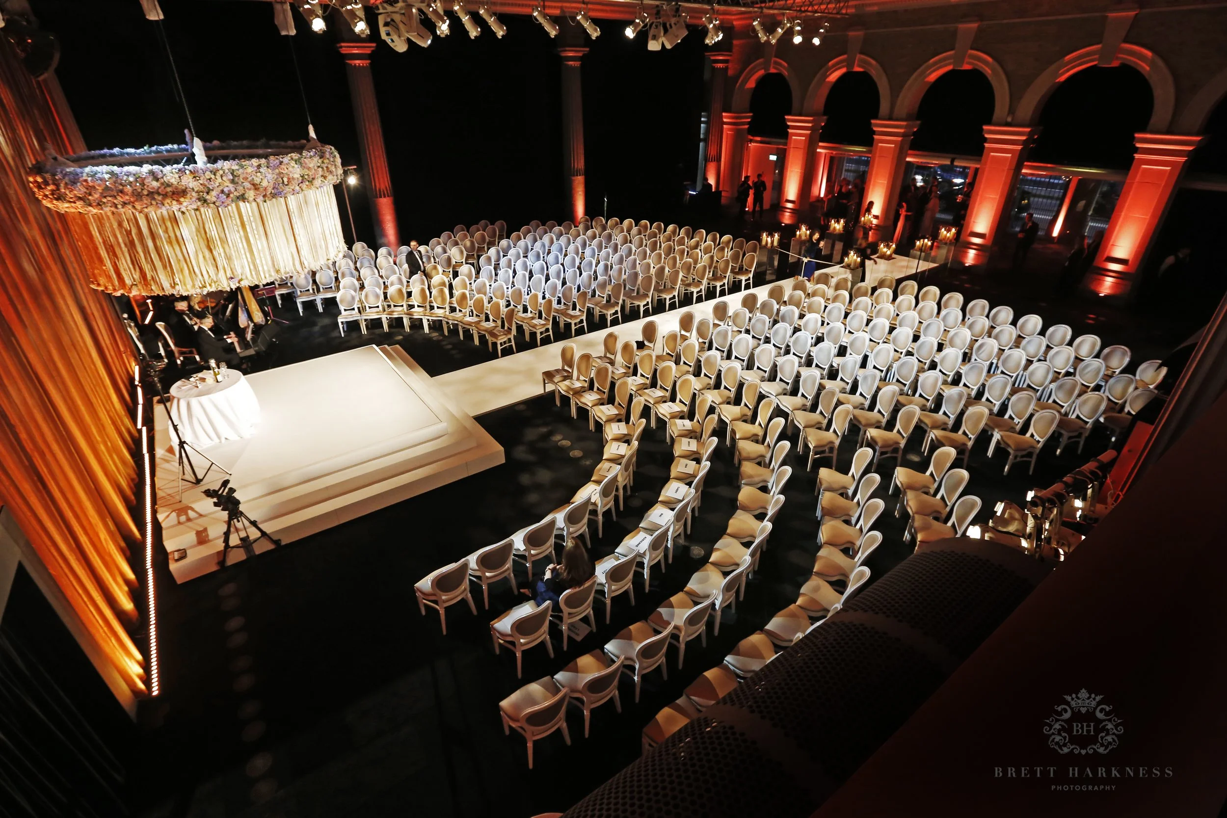 An elegant indoor event space set for a wedding with rows of white chairs arranged in front of a stage, a large floral chandelier hanging above, warm orange lighting on the walls, and a few people preparing for the ceremony.