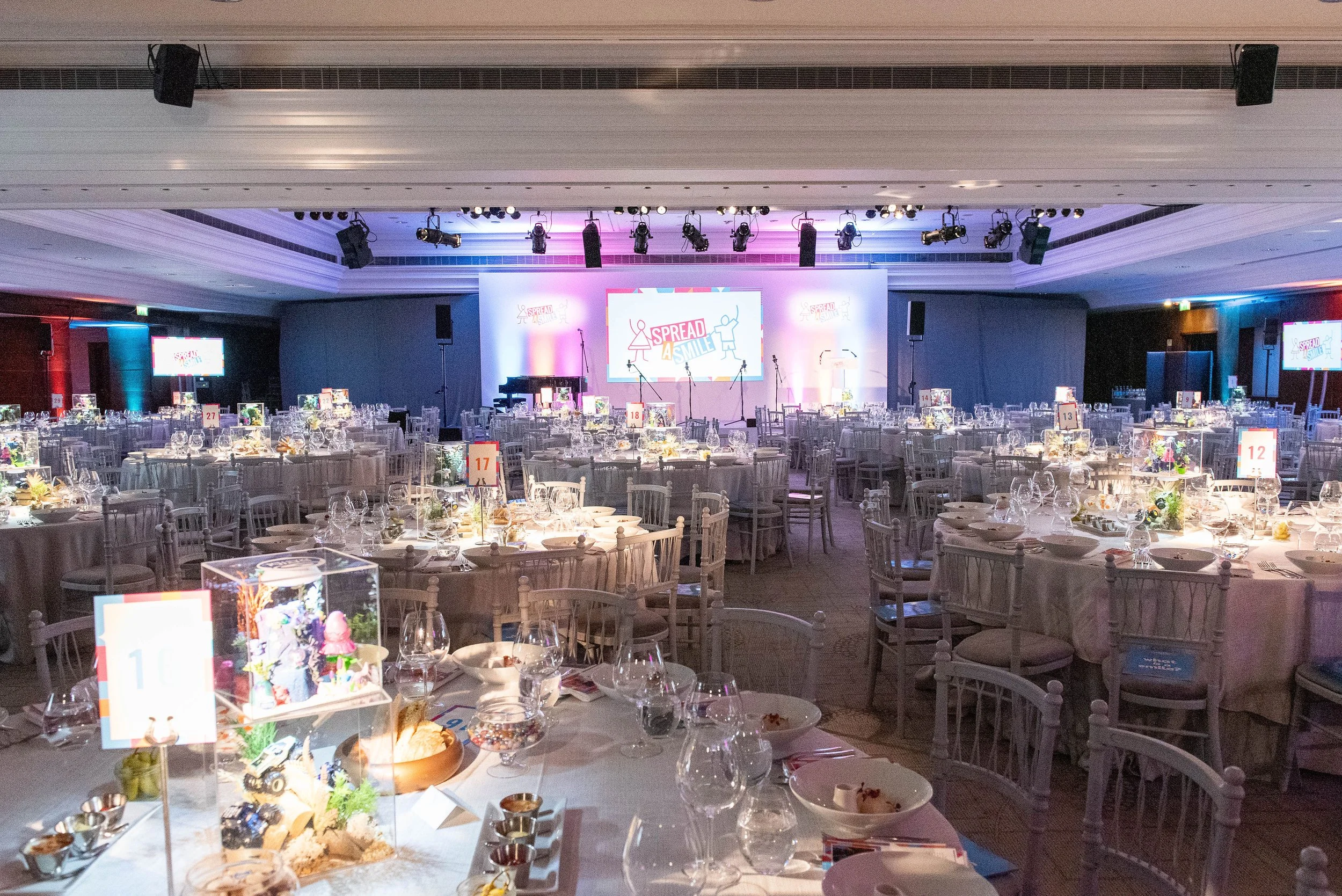 Event space with round tables set with glassware, plates, and napkins, a stage with large screens displaying 'Spread a Smile' in the background, and colorful lighting.
