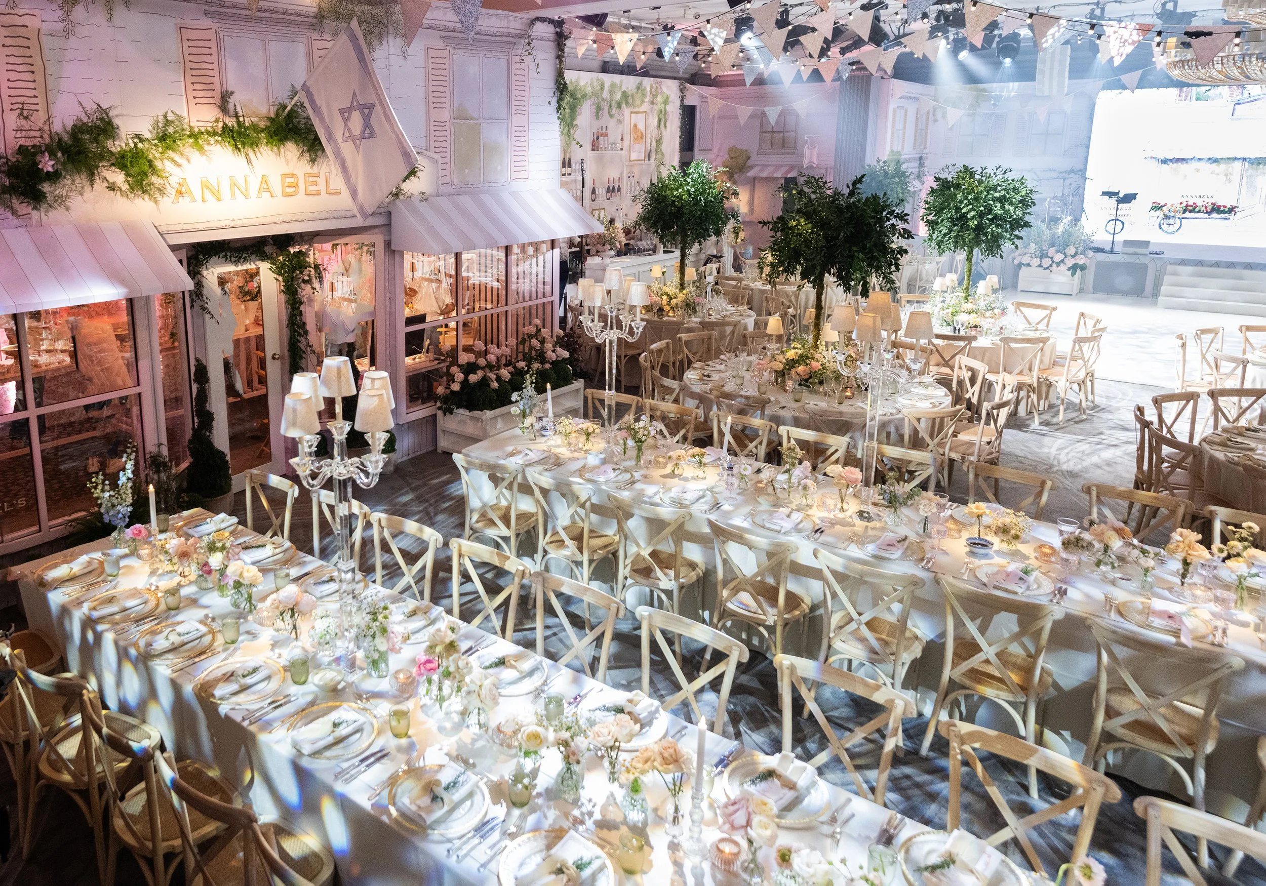 Decorated indoor event space with round tables set with white tablecloths, floral centerpieces, candles, and place settings. There are potted trees, chandeliers, and string banners hanging from the ceiling at Grosvenor House on Park Lane.