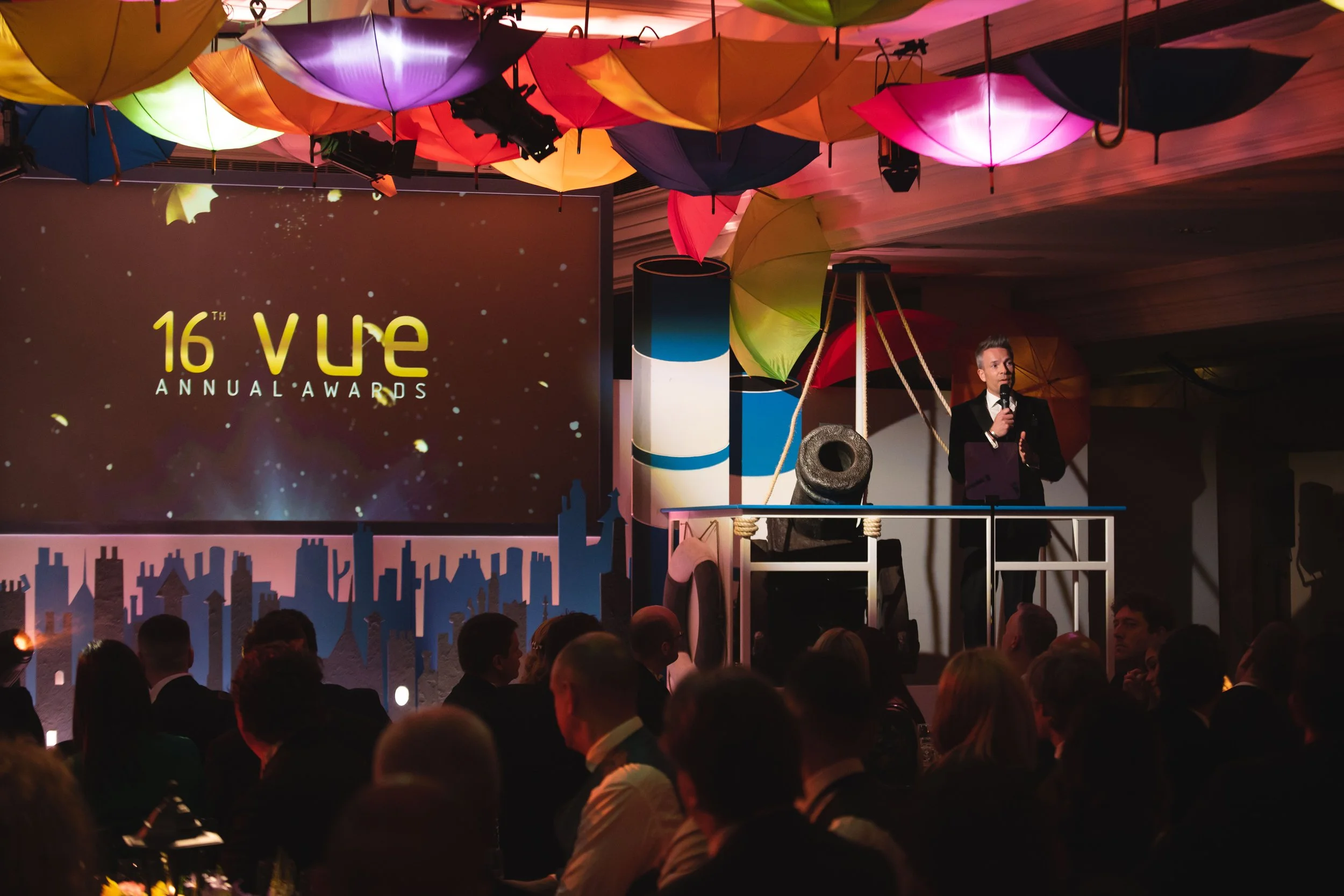 A stage with a large screen displaying '16th VUE Annual Awards'. There are several colorful umbrellas hanging upside down above the stage. A man in a black suit and white shirt is speaking into a microphone on the stage. The audience is visible in th
