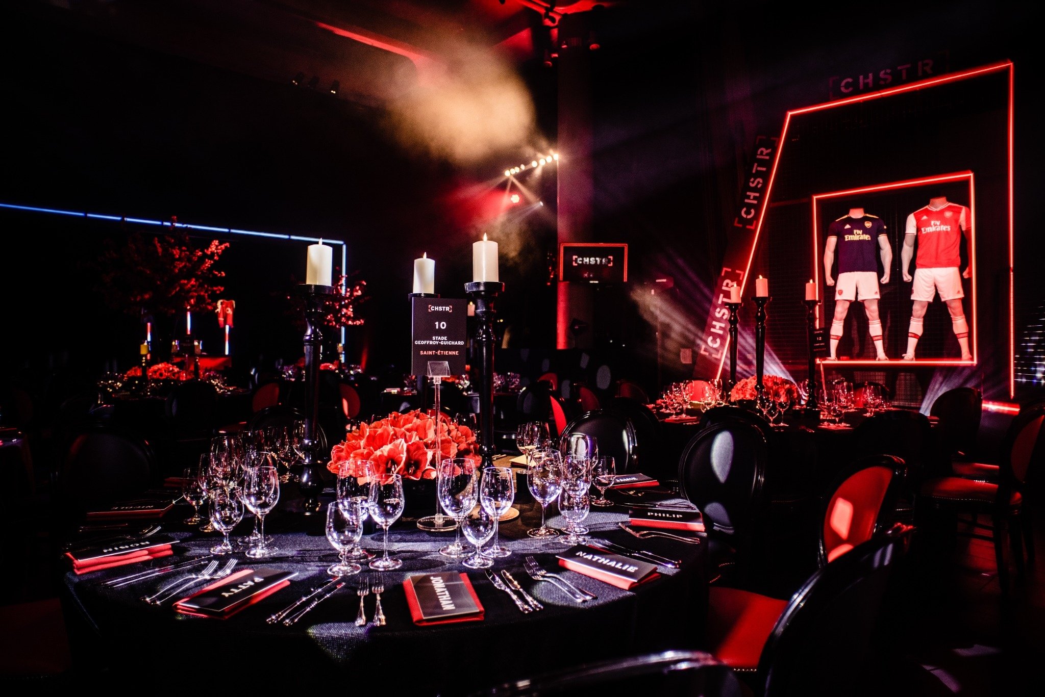 A banquet hall decorated in black and red for a sports-themed event, featuring tables set with glassware, flowers, and place cards, with mannequins dressed in football jerseys on a stage in the background, and candles on the tables.