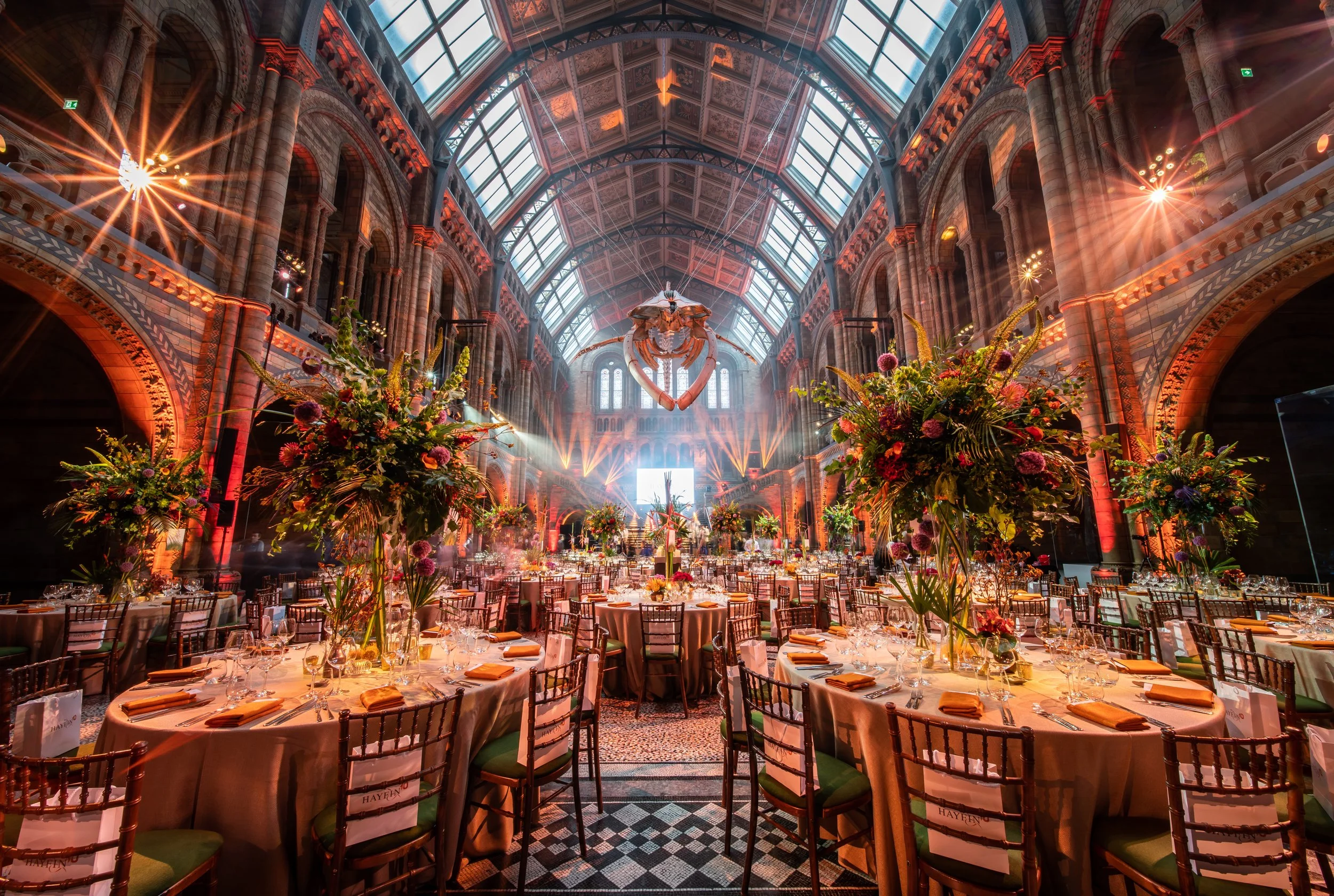 The Natural History Museum glass and stone building with high vaulted glass ceiling, decorated with large floral arrangements, set up for a corporate annual anniversary with round tables, chairs, and tableware.