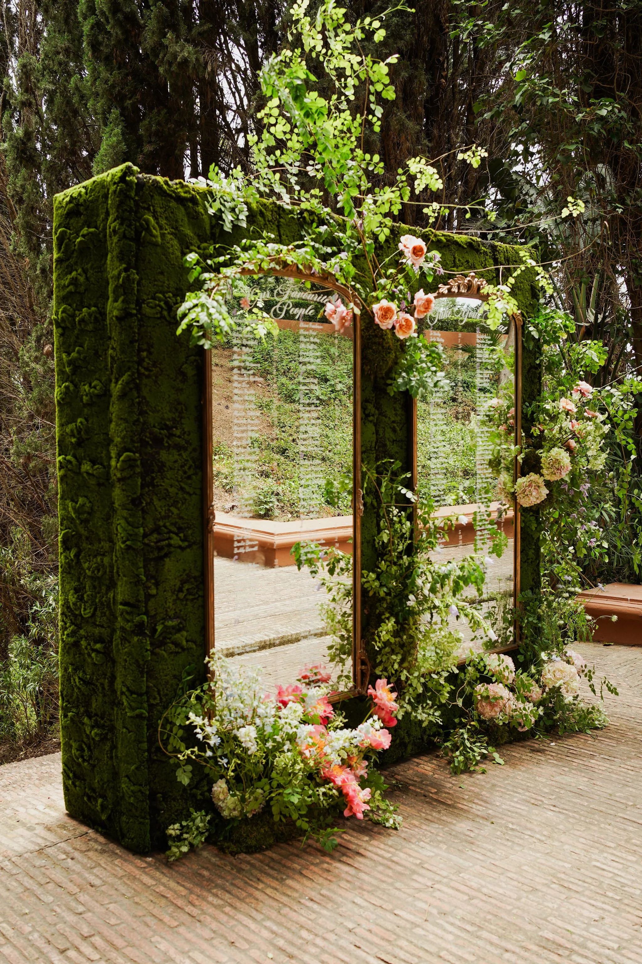 A decorative mirror with a moss-covered frame, adorned with pink and white flowers and green foliage, is placed outdoors against a backdrop of trees and plants at Anantara Villa Padierna Resort in Marbella