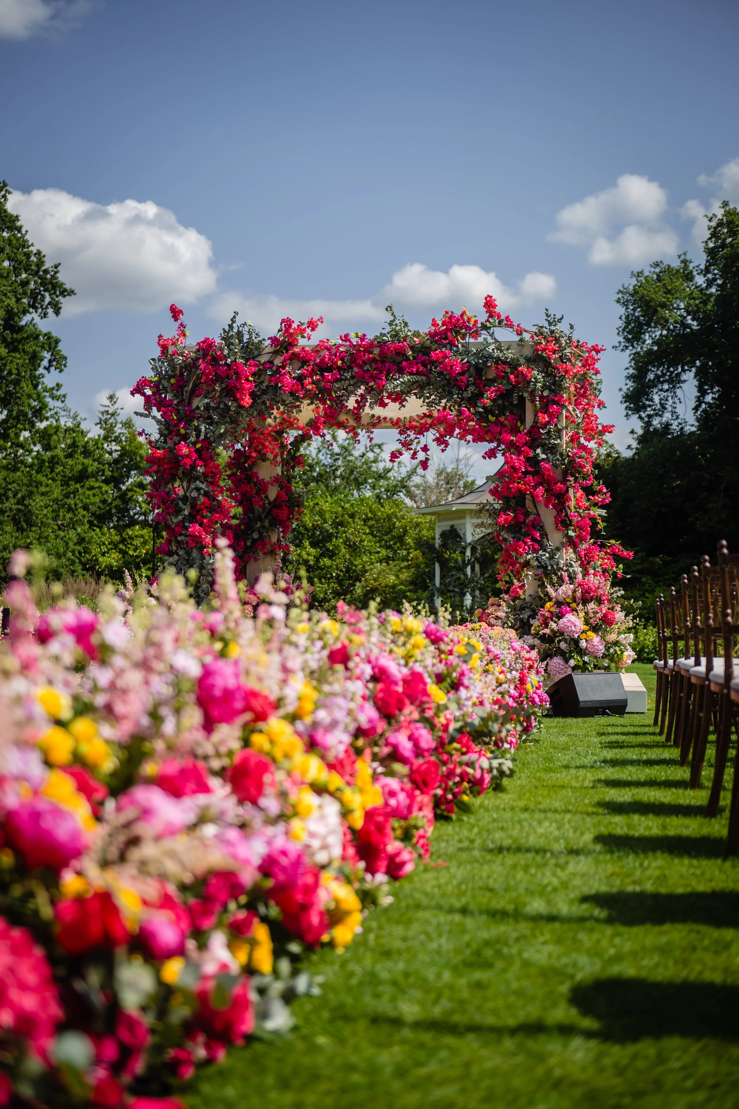 A garden with a floral arch decorated with pink and red flowers, surrounded by colorful flowers on the ground and chairs set up for an outdoor event. Fairmont Windsor Park'.