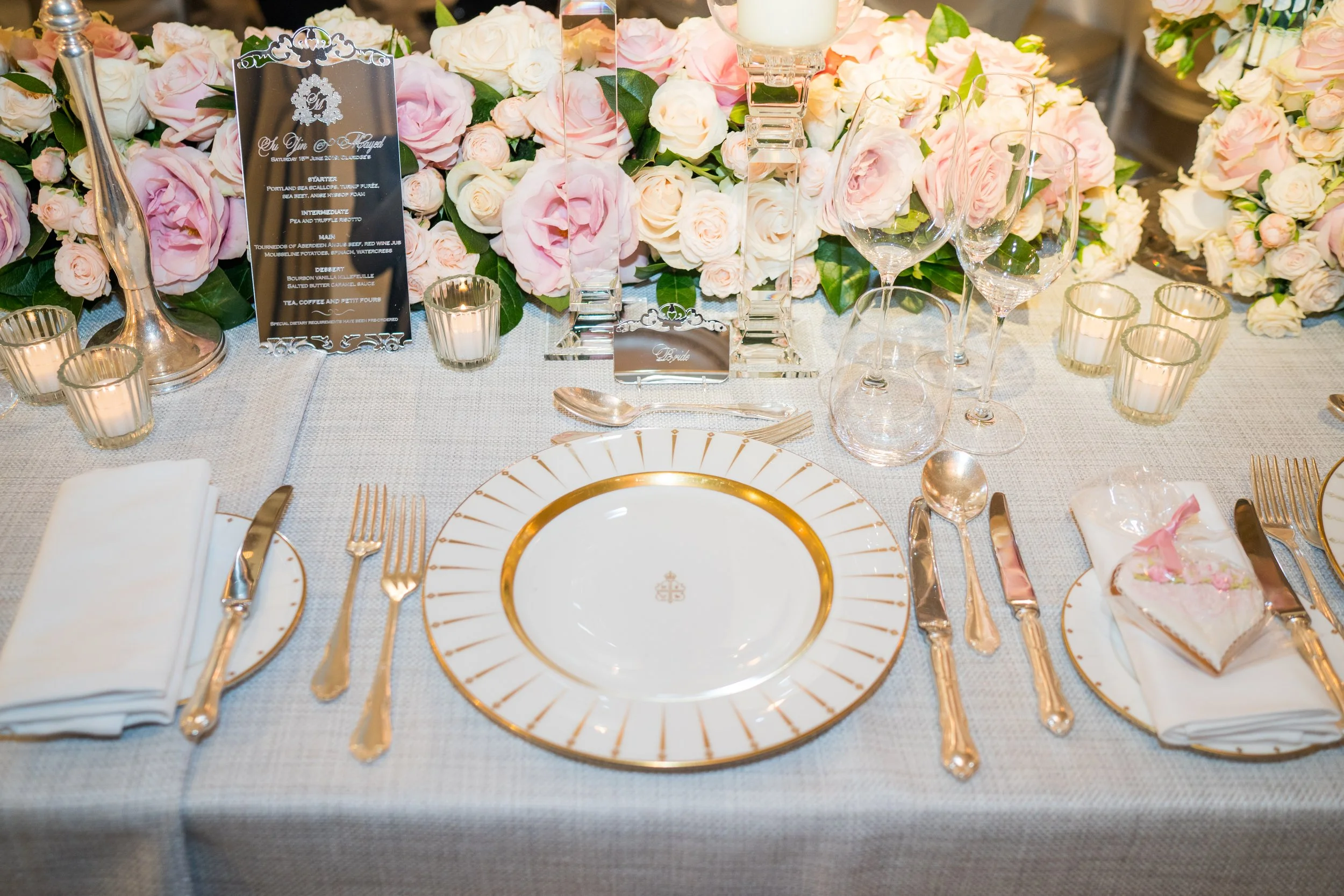 Elegant wedding table setup with white and pink roses, glassware, gold-accented plates, cutlery, and a menu card.
