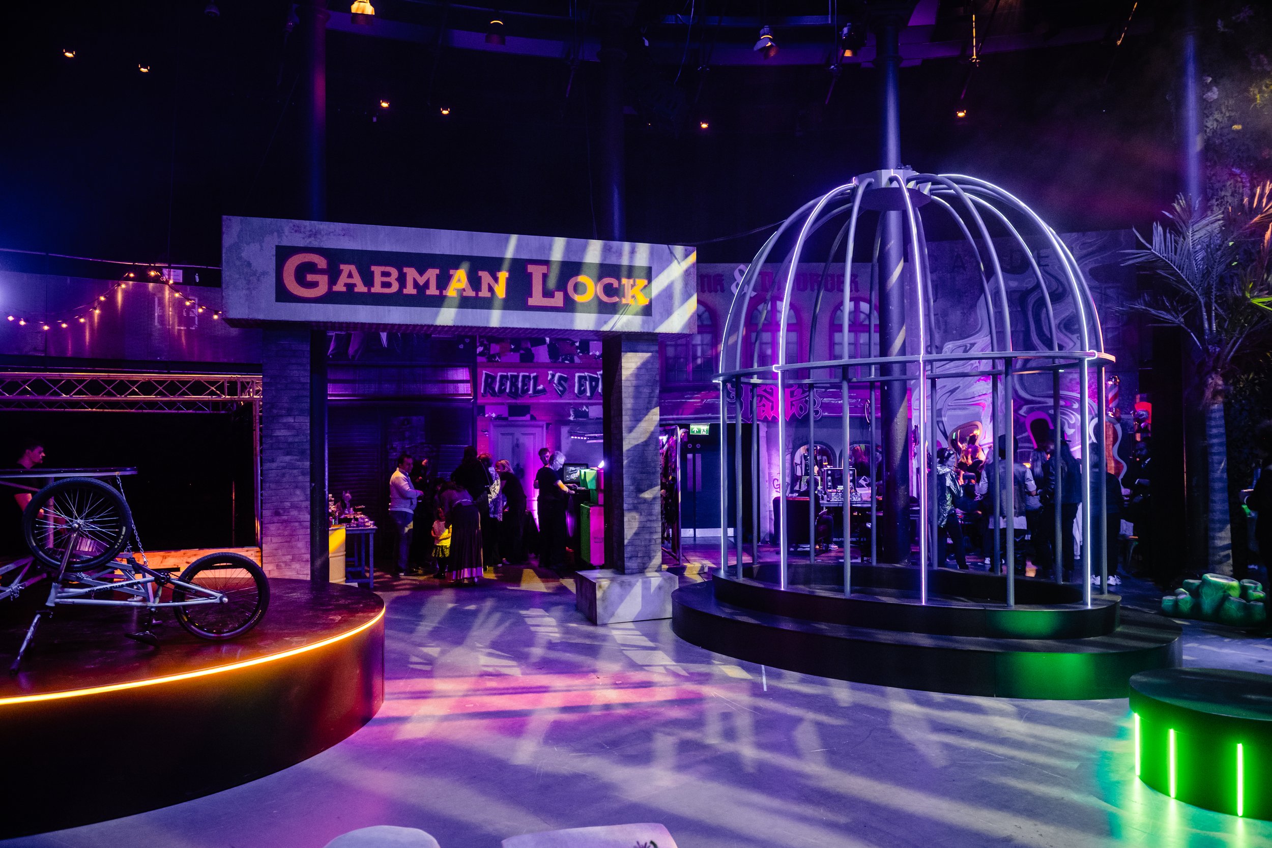 Colorful Roundhouse venue with neon lights, a sign reading 'GABMAN LOCK', and various amusement rides and attractions, with groups of people enjoying the space at a Bar Mitzvah 