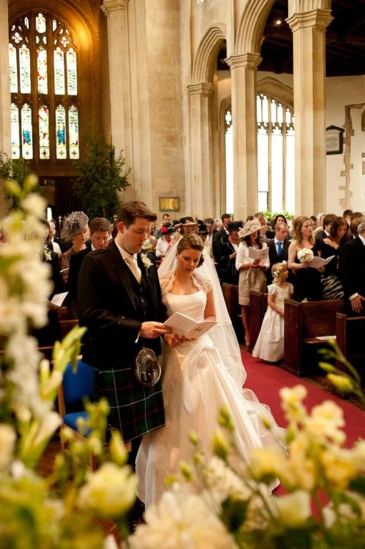 A wedding ceremony inside a church with a bride and groom standing together, reading or listening to a program, surrounded by guests.