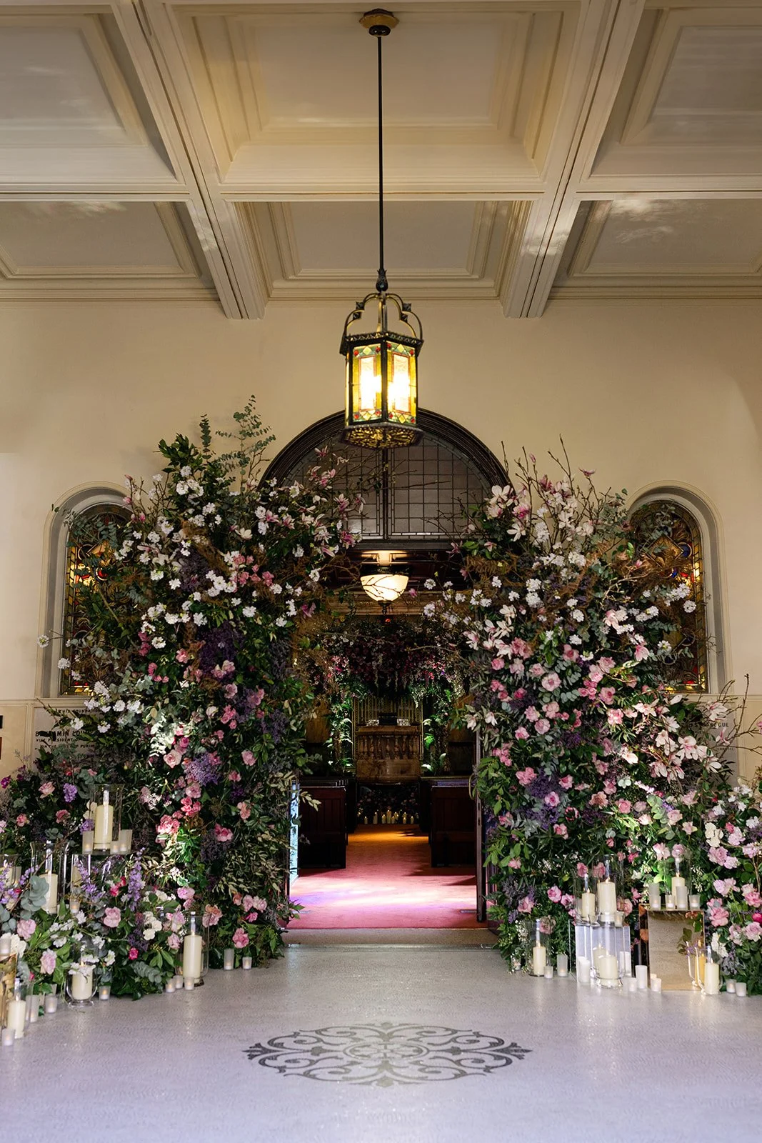 Elegant church entrance decorated with abundant pink, purple, and white flowers and candles, with stained glass windows and a hanging lantern.