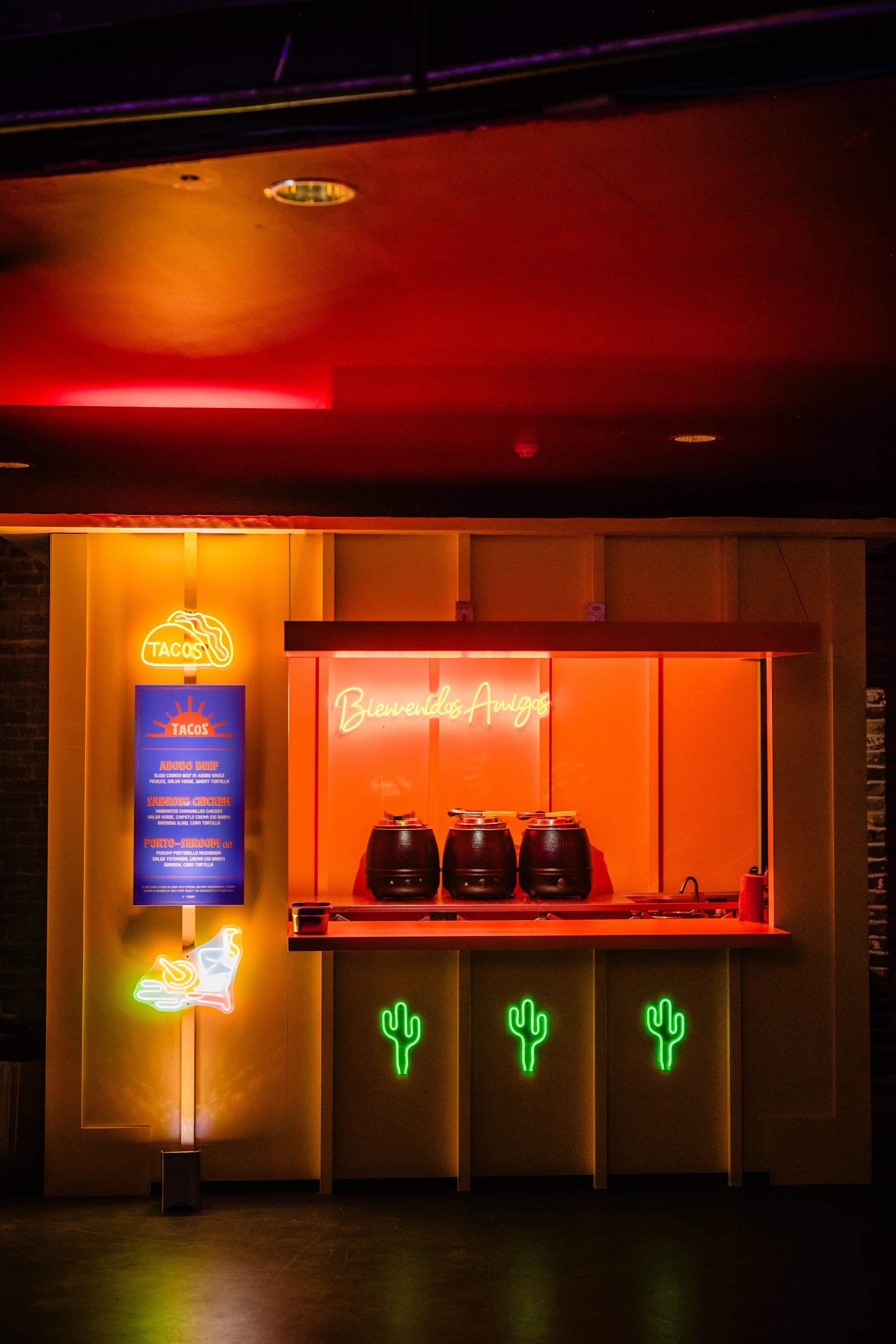 Neon sign with a taco and cactus themed decor, including a welcoming message in Spanish, beneath three black containers on a counter, with additional neon signs advertising tacos and featuring cactus and hand graphics.