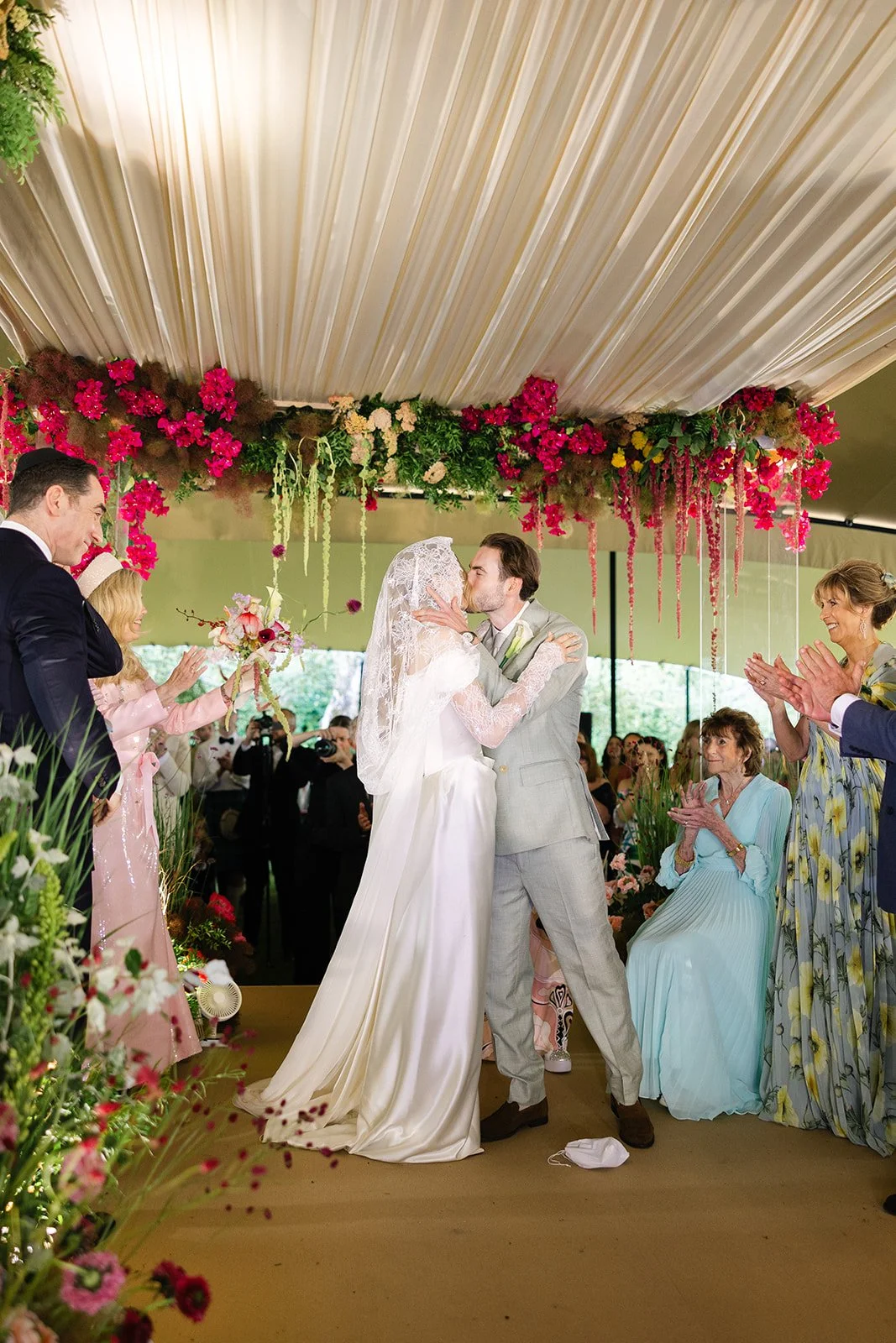 A wedding ceremony with a bride and groom kissing under a floral and greenery chuppah, surrounded by guests clapping in a decorated tent.