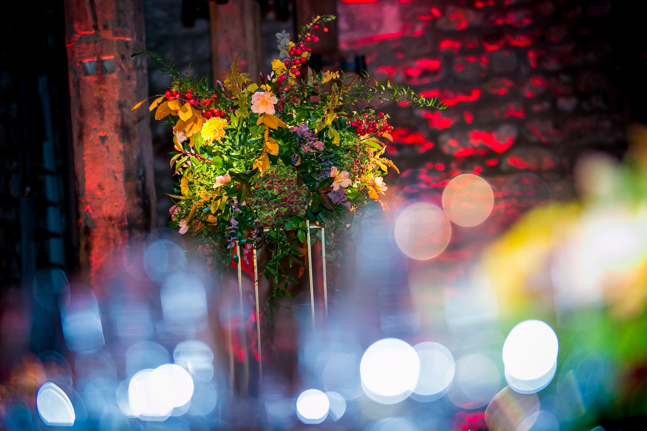 A bouquet of colorful flowers and red berries on a stand against a dark rustic wall, illuminated with warm lighting creating a bokeh effect with colorful circular light spots at the Tower of London