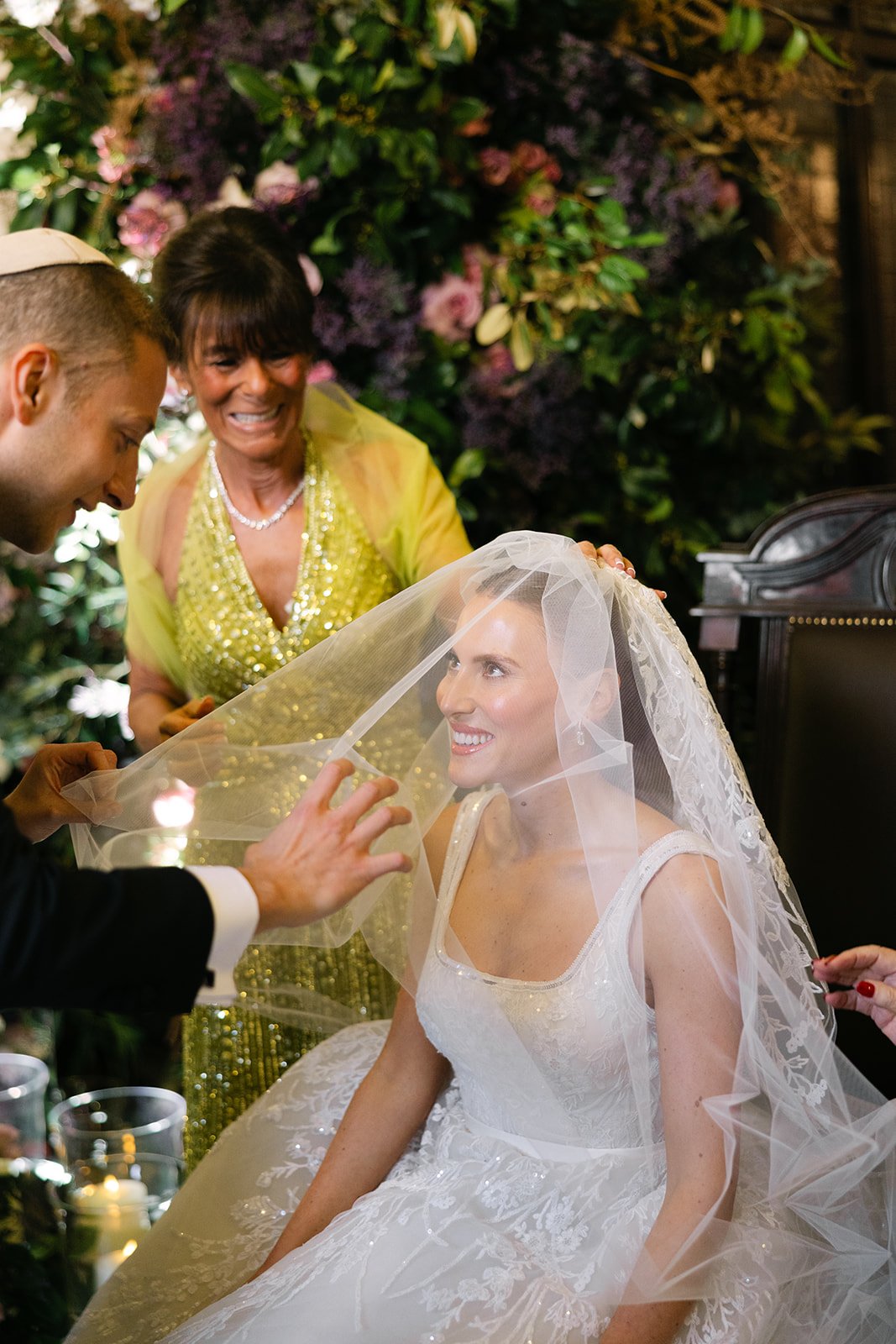 A bride in a white wedding dress is smiling as her veil is being adjusted by a man, with a woman in a bright yellow dress and a pearl necklace smiling behind her, in a floral setting. Jewish wedding