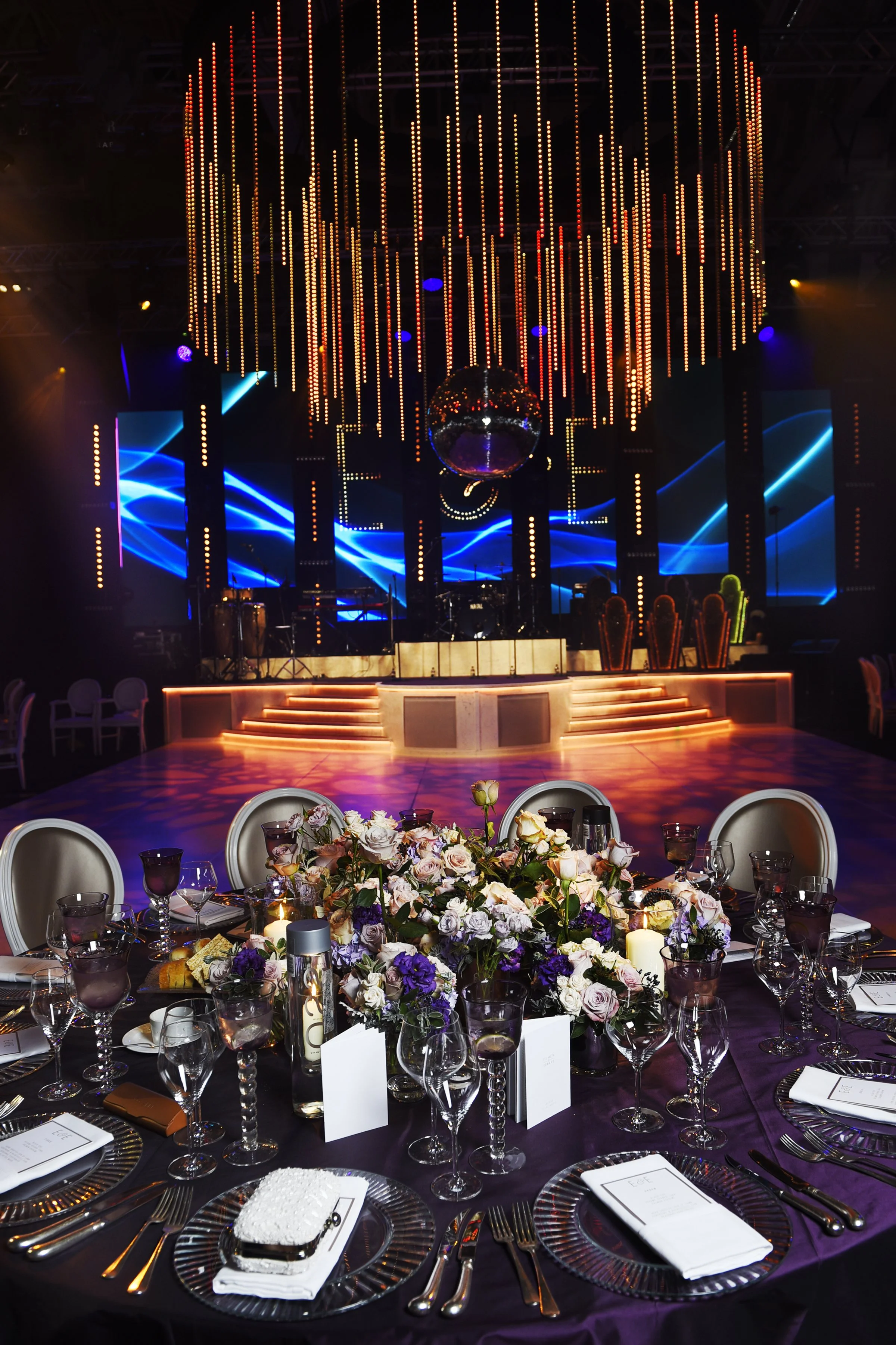A decorated banquet table with a floral centerpiece, candles, and place settings, set in front of a stage with blue lights, a disco ball, and vertical light strips in a dark venue.