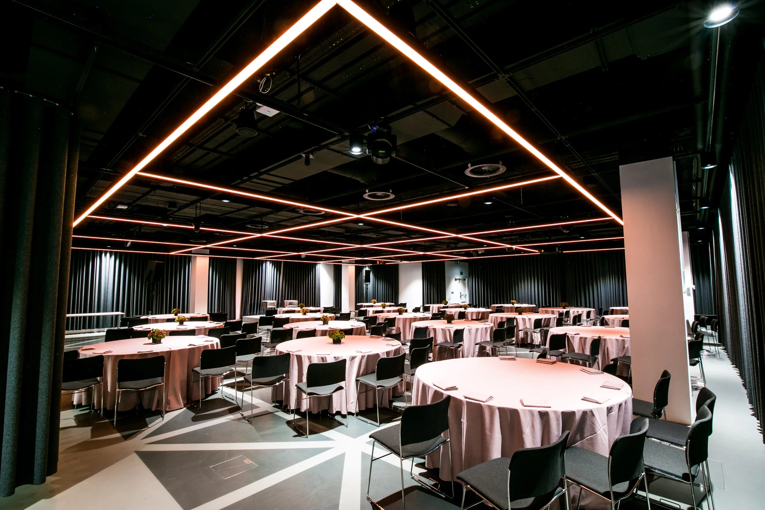 A modern event space with round tables covered in pink tablecloths, each decorated with small floral centerpieces. The ceiling features geometric light fixtures emitting a soft glow, and the room has dark curtains along the walls.