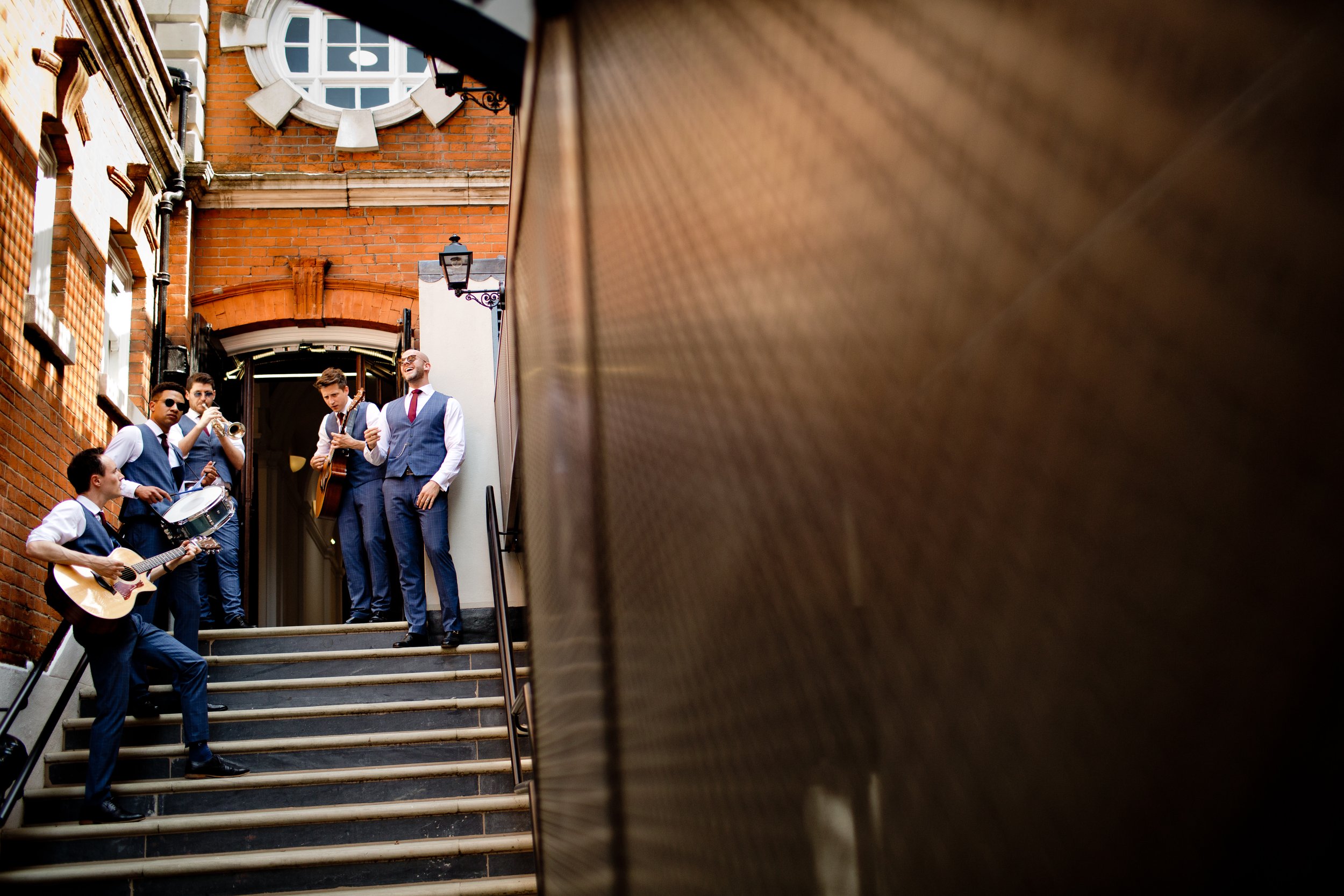 A group of five men in vests and ties, some holding musical instruments, standing on stairs outside a brick building with a round window, possibly preparing to perform or during a musical event.