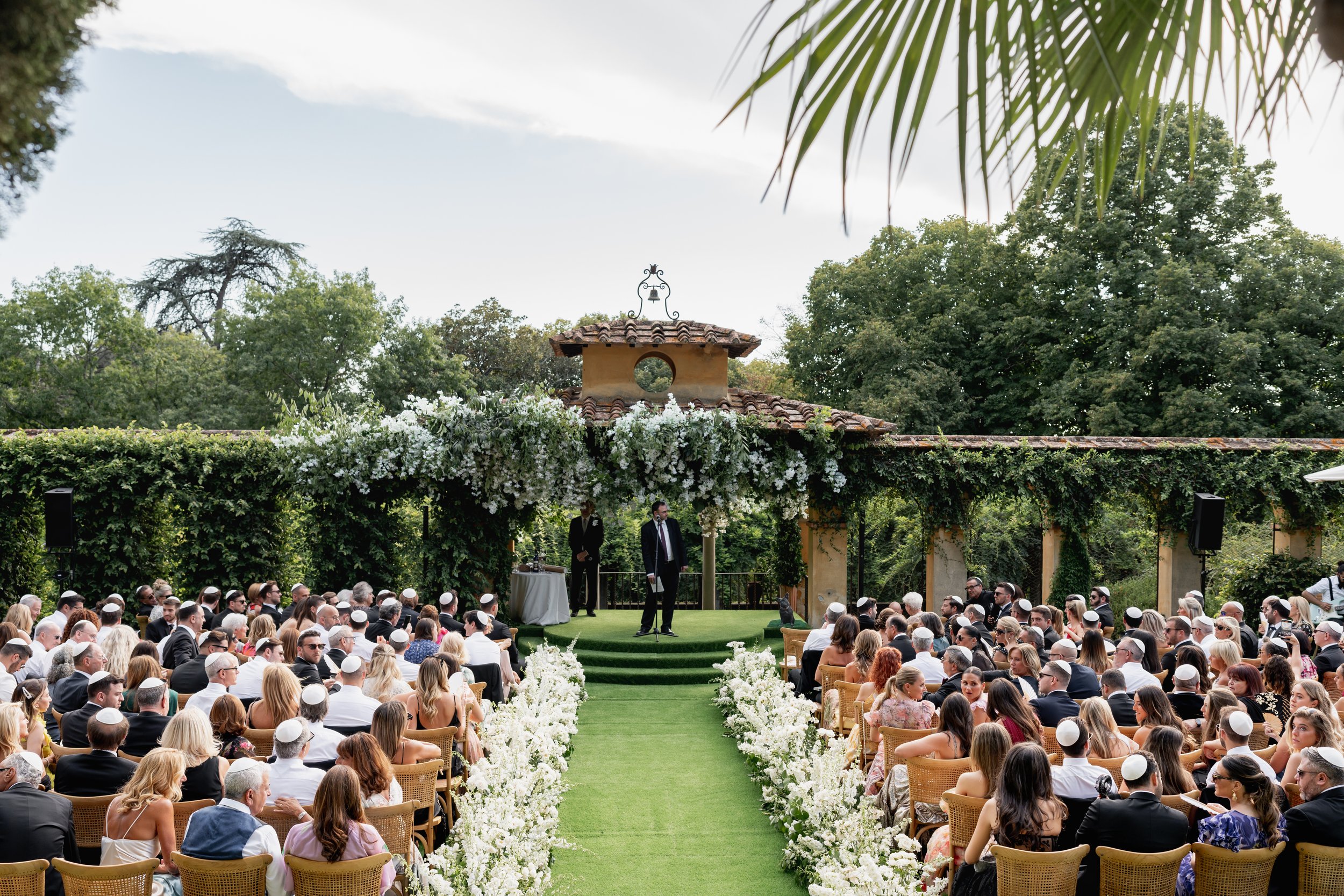 A large outdoor wedding ceremony with guests seated on wicker chairs under a decorated canopy, surrounded by greenery and trees at Villa Palmieri in Florence