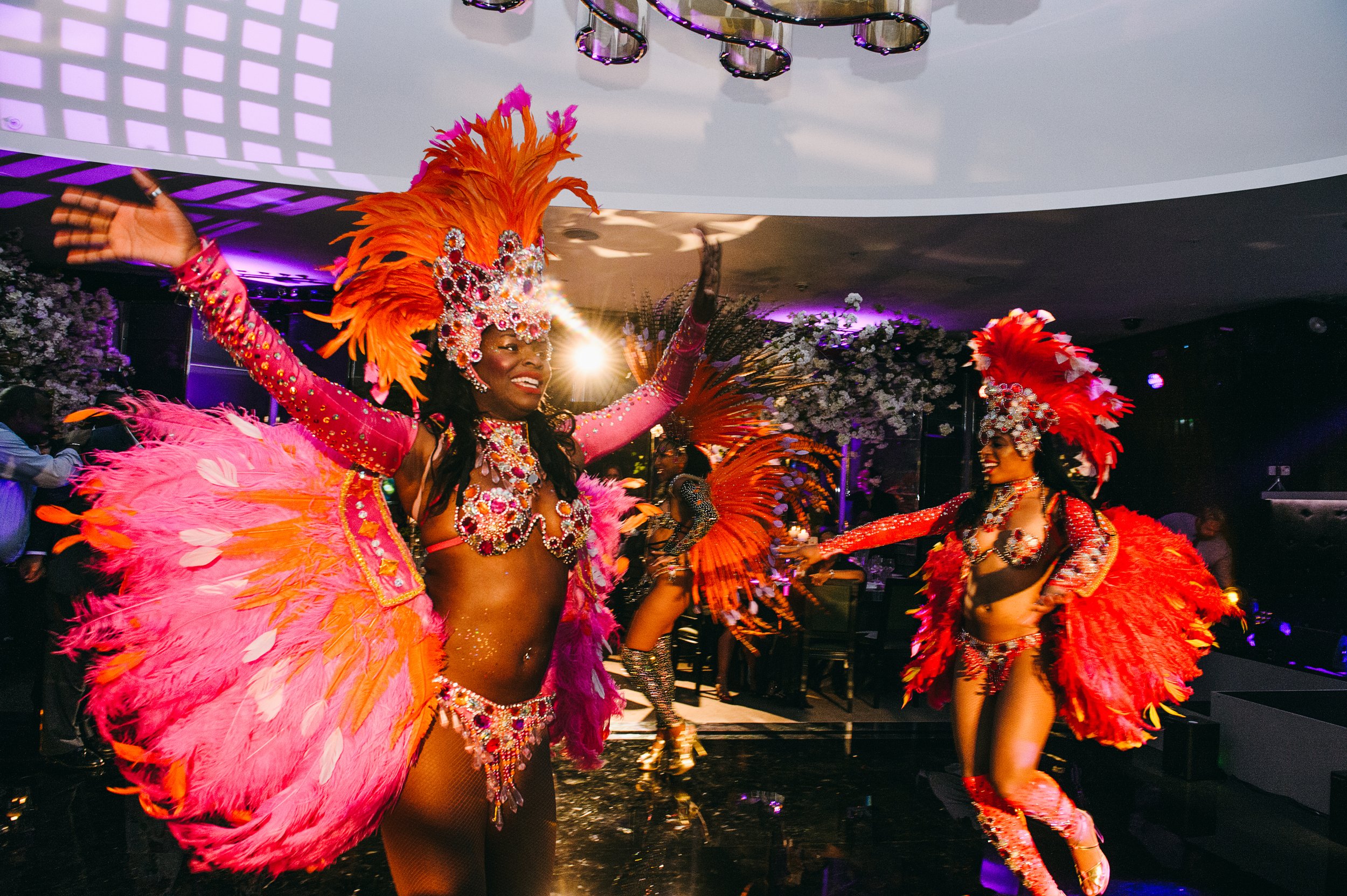 Women dressed in colorful carnival costumes dancing in a party or celebration setting with purple and white lighting.
