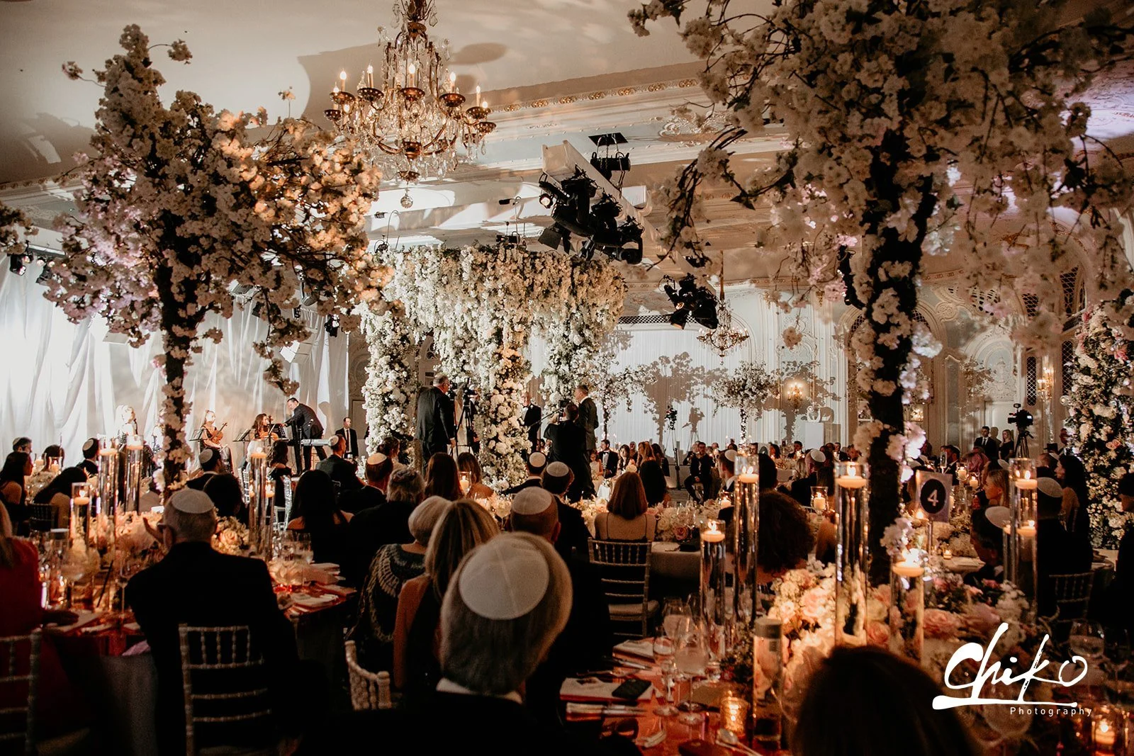 A large banquet hall decorated for a formal event or wedding, with tall floral arrangements of white and pink flowers, a chandelier, and a stage with musicians performing. The room is filled with seated guests, many wearing traditional Jewish head co