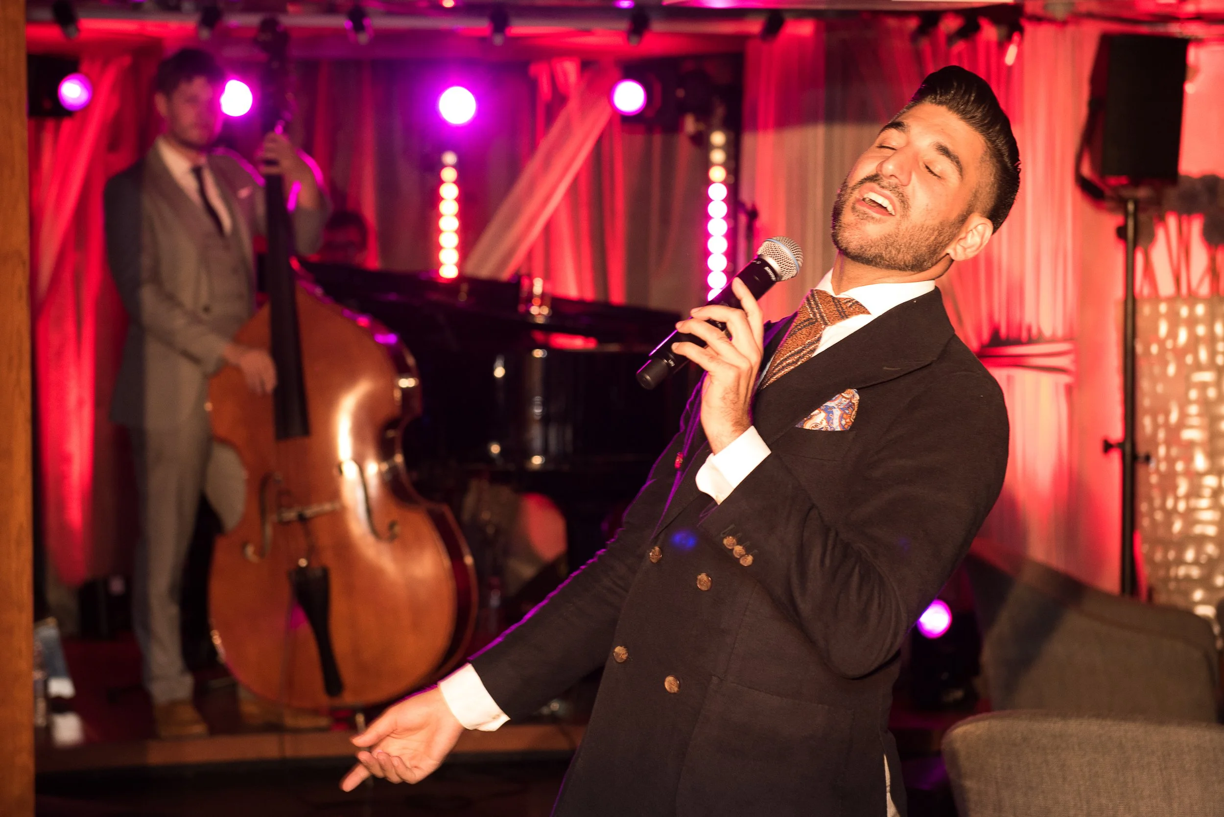 A man in a black suit and patterned tie singing into a microphone on stage, with a musical band and upright bass in the background, during a live performance in a dimly lit venue with pink and purple lighting.
