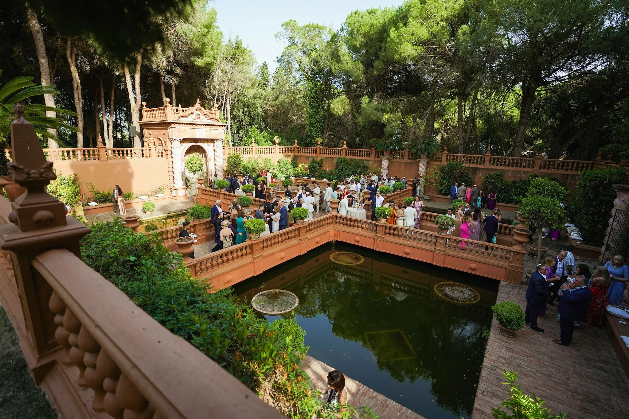 A gathering of people at an outdoor event in a garden courtyard with a large pond, surrounded by greenery and tall trees, with a decorative archway and ornate brick railing at Anantara Villa Padierna Resort in Marbella