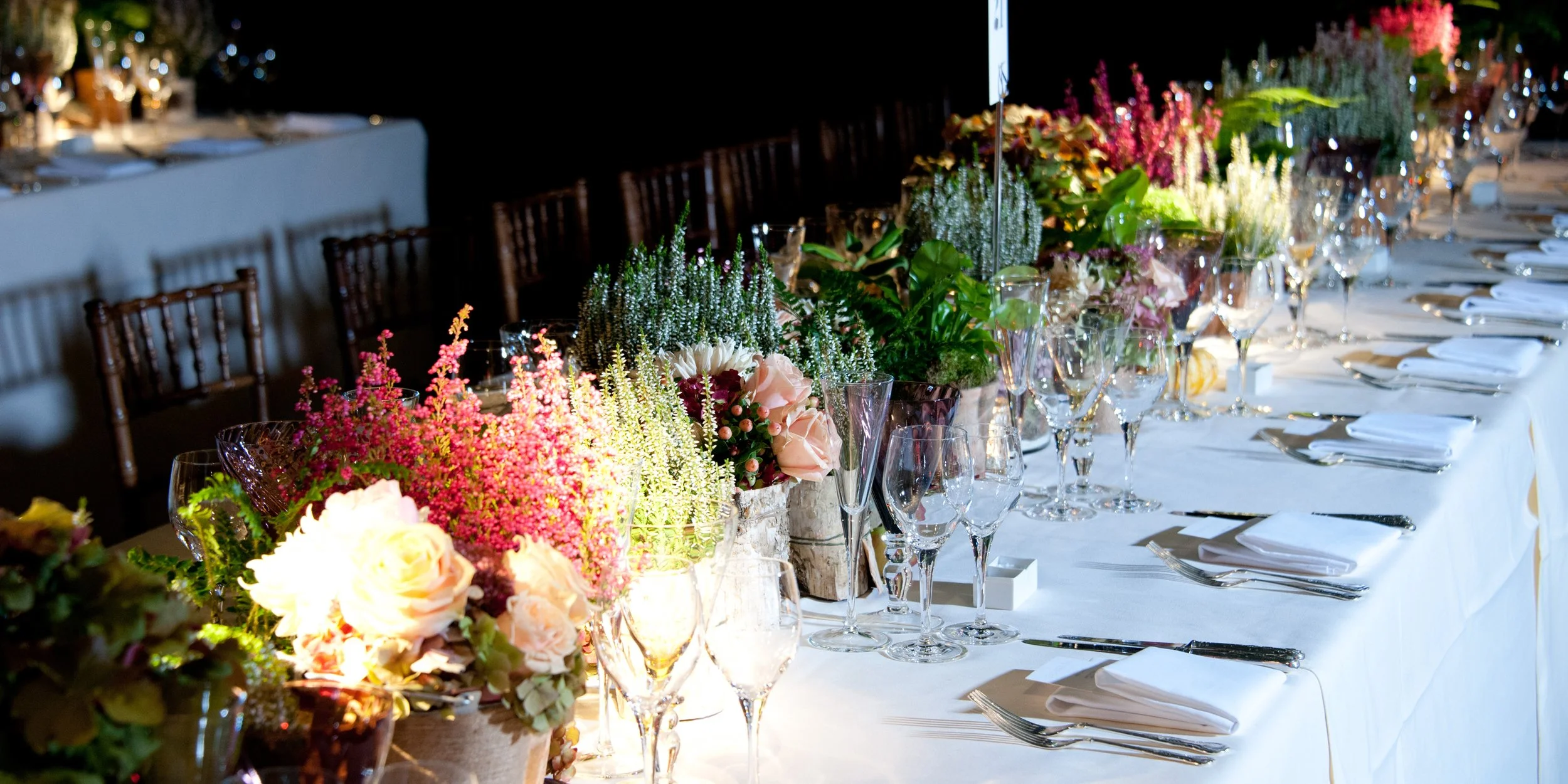 A long banquet table with a white tablecloth, set with multiple wine glasses, silverware, and white napkins, decorated with colorful flower arrangements including roses and other blooms.