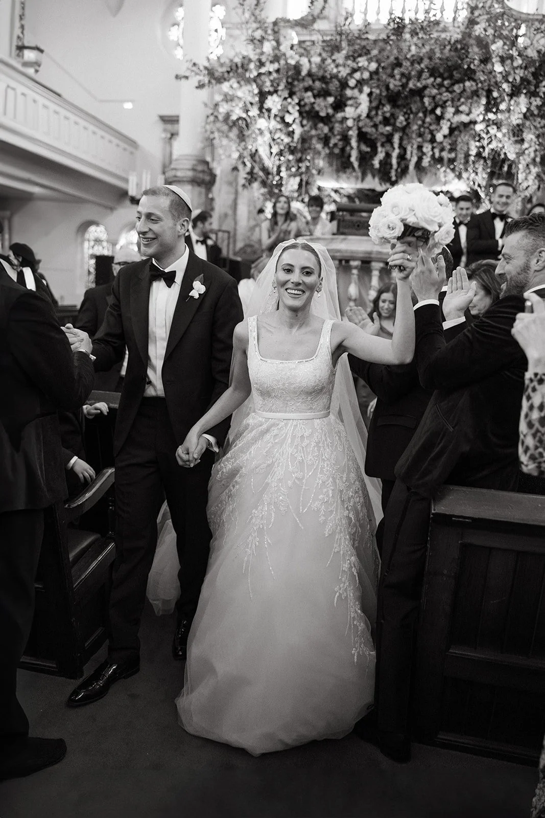 A bride holding a bouquet of roses and smiling, walking down the aisle with a groom at her side, surrounded by guests in a church decorated with flowers. Jewish wedding