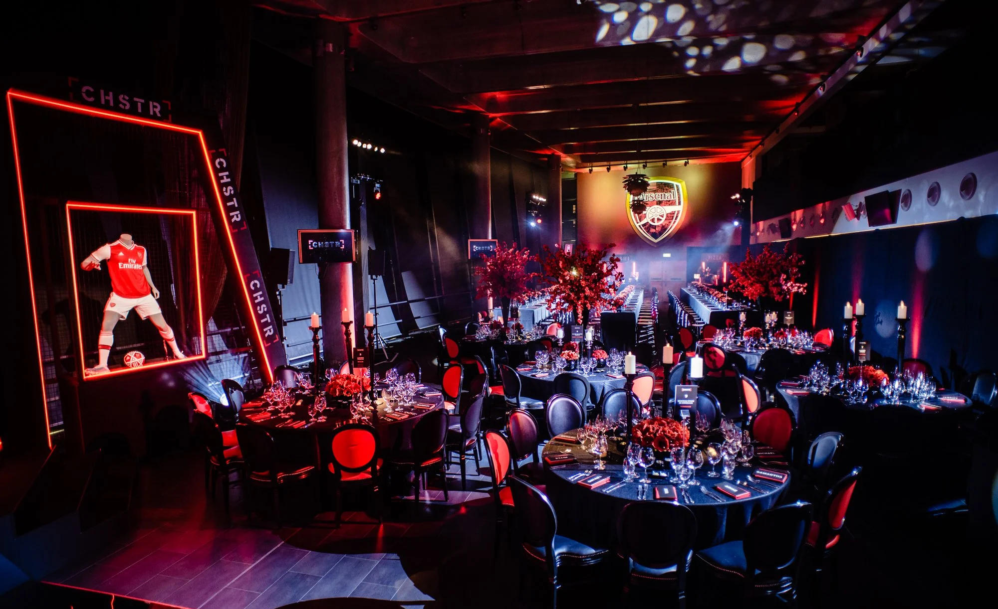 Event hall decorated with Arsenal Football Club theme, featuring tables with black and red chairs, floral arrangements, candles, and large displays of Arsenal team jerseys and logo.