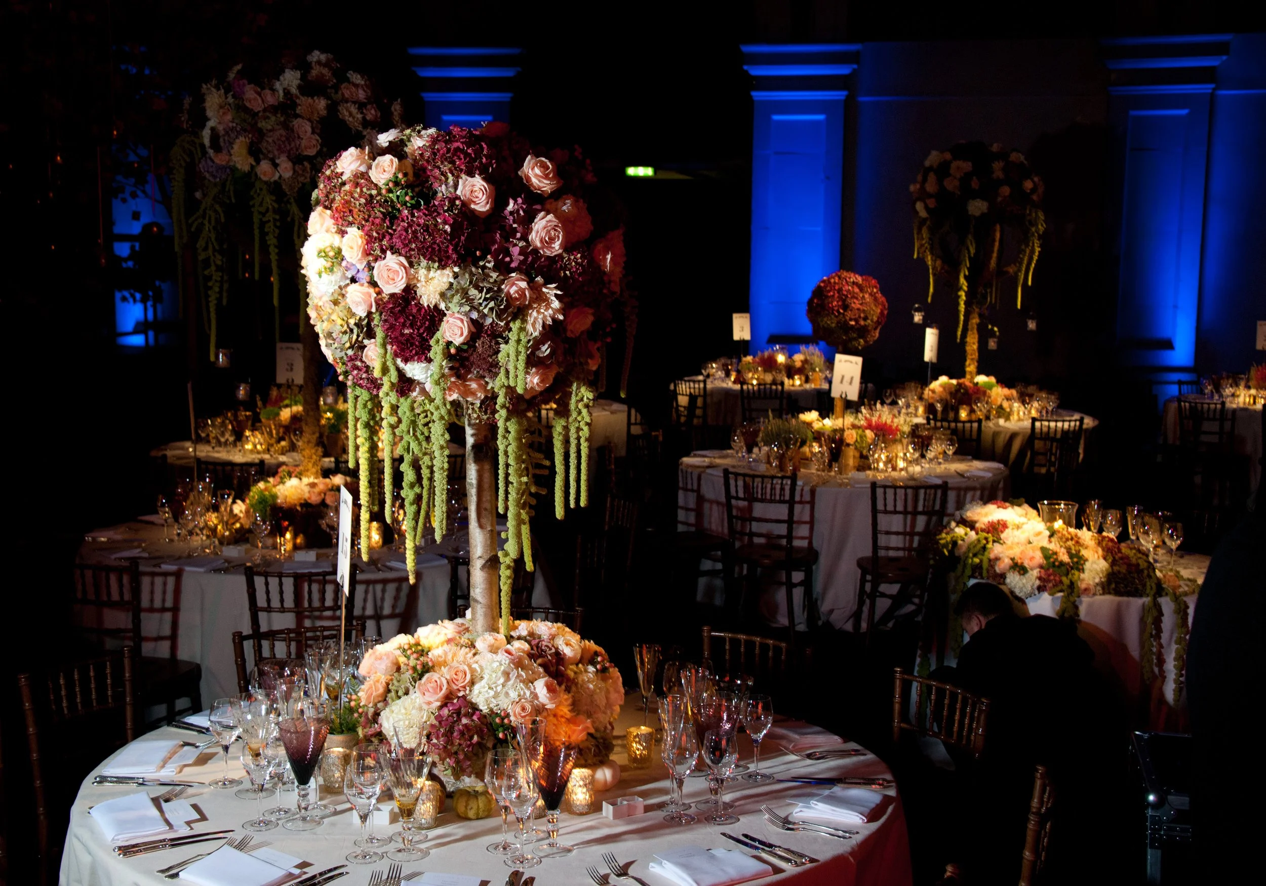 Elegant banquet hall decorated with large floral arrangements featuring pink roses, purple hydrangeas, and cascading green amaranthus, illuminated with warm lighting and blue uplighting on the walls, set with round tables, candles, wine glasses, and 