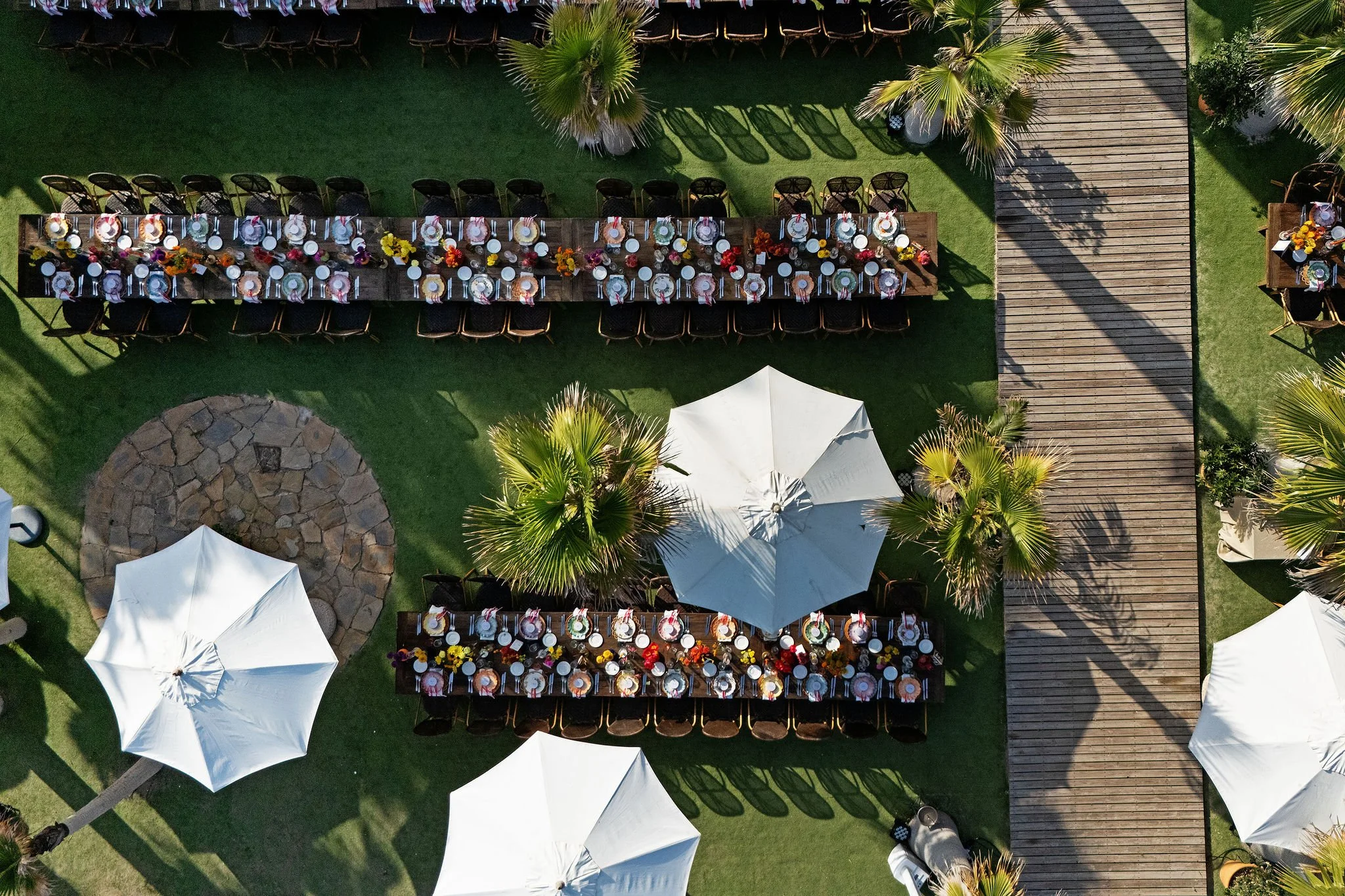Aerial view of a large outdoor dining setup on a lawn with a long wooden table decorated with colorful flowers and place settings, shaded by white umbrellas, and surrounded by palm trees and a wooden deck at Anantara Villa Padierna Resort in Marbella