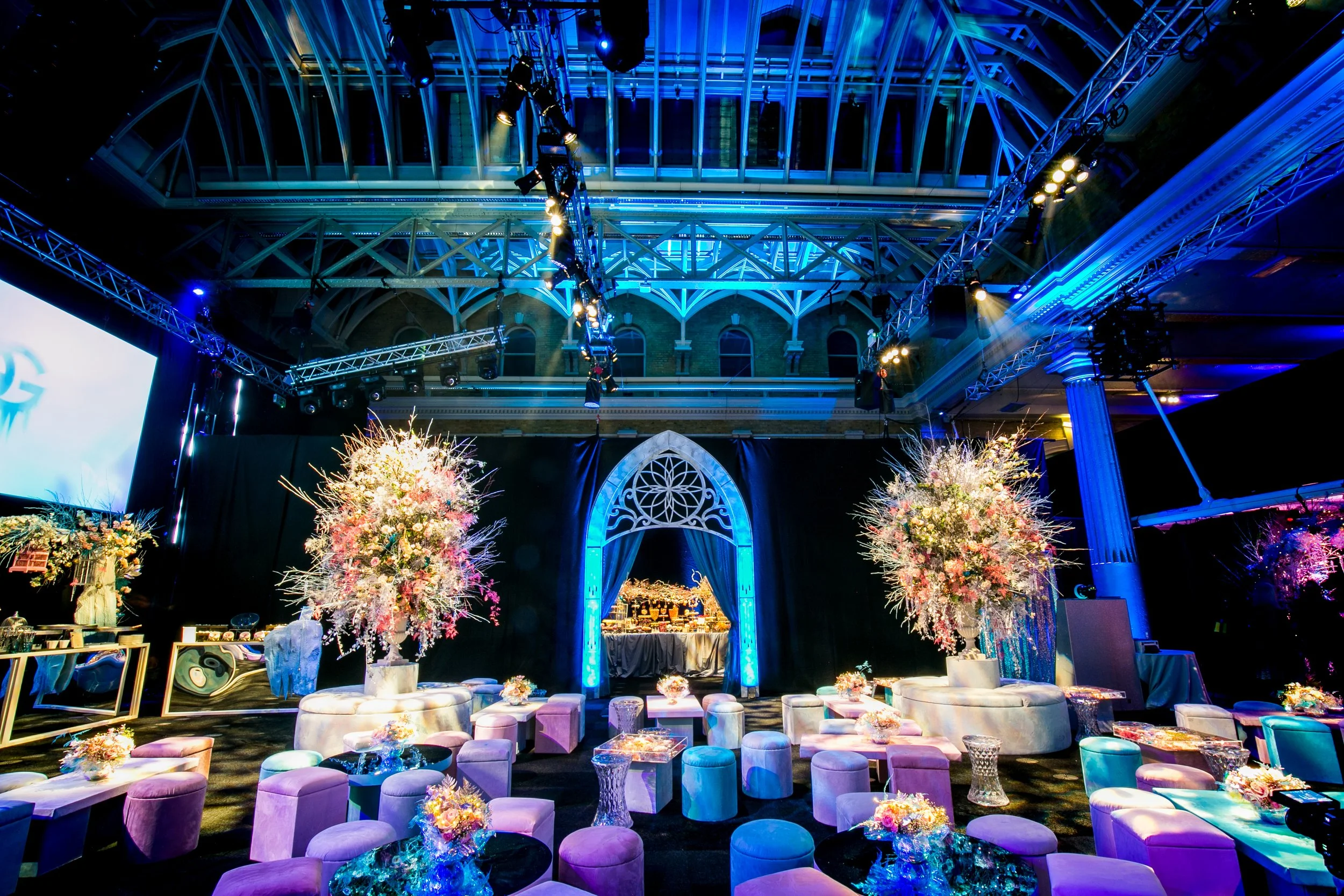 Illuminated event space with purple, pink, and blue lighting, floral arrangements, small tables, and seating in a large architectural venue with a glass ceiling. ICE PALACE BAT MITZVAH at Old Billingsgate 