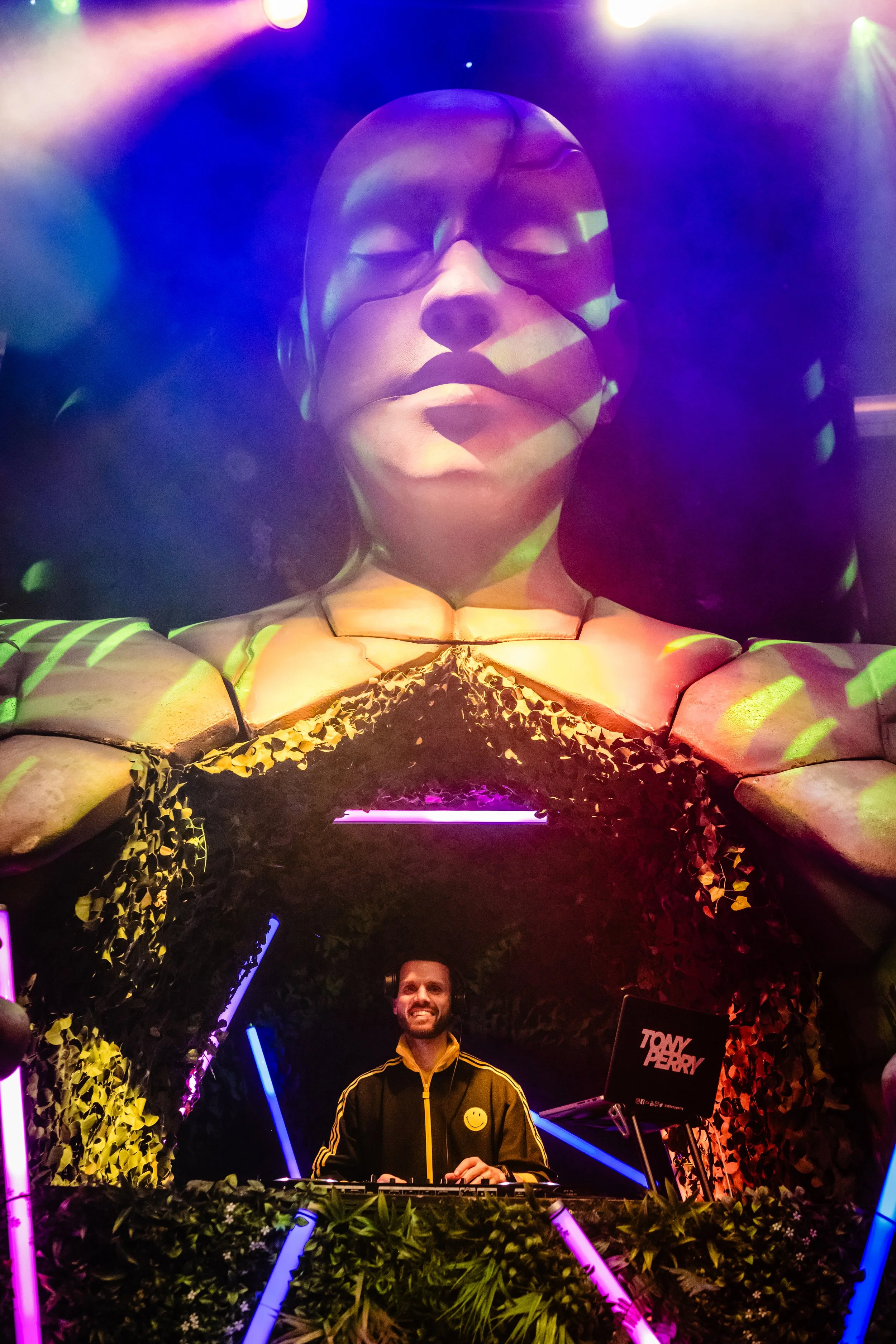 A DJ with a yellow smiley face on his jacket is playing music in a colorful, vibrant nightclub with neon lights, a large face sculpture with sunglasses above, and tropical-themed decorations at a Bar Mitzvah 