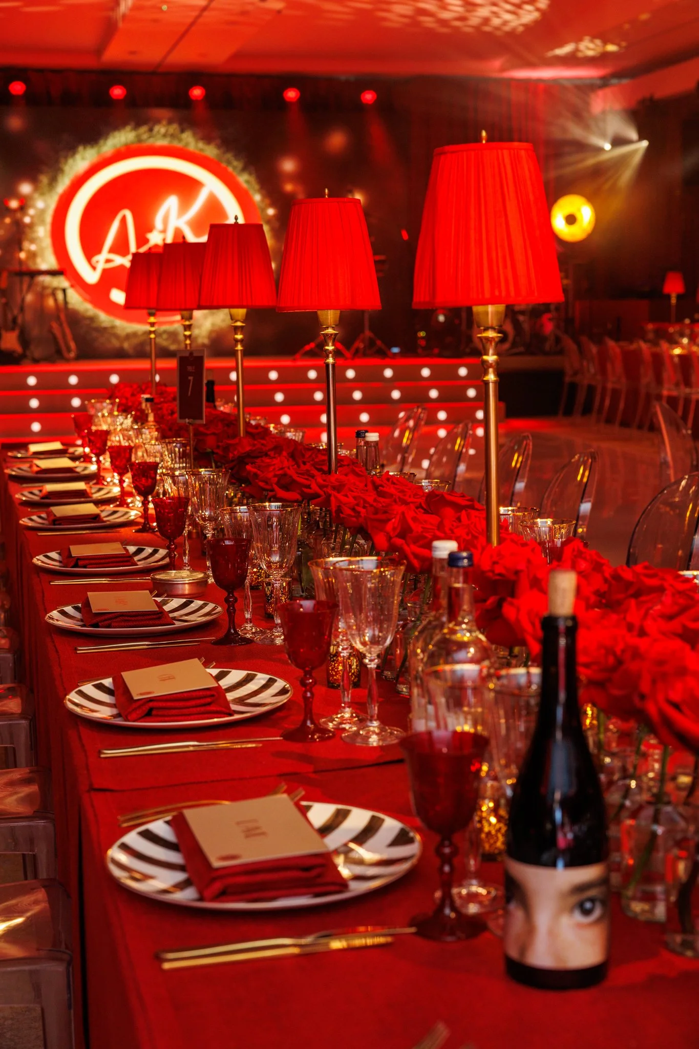 A decorated dining table for a celebration or event with red tablecloths, red lampshades, glasses, plates with zebra print, and a centerpiece of red roses. In the background, there is a stage with a large illuminated logo at at The Grove 