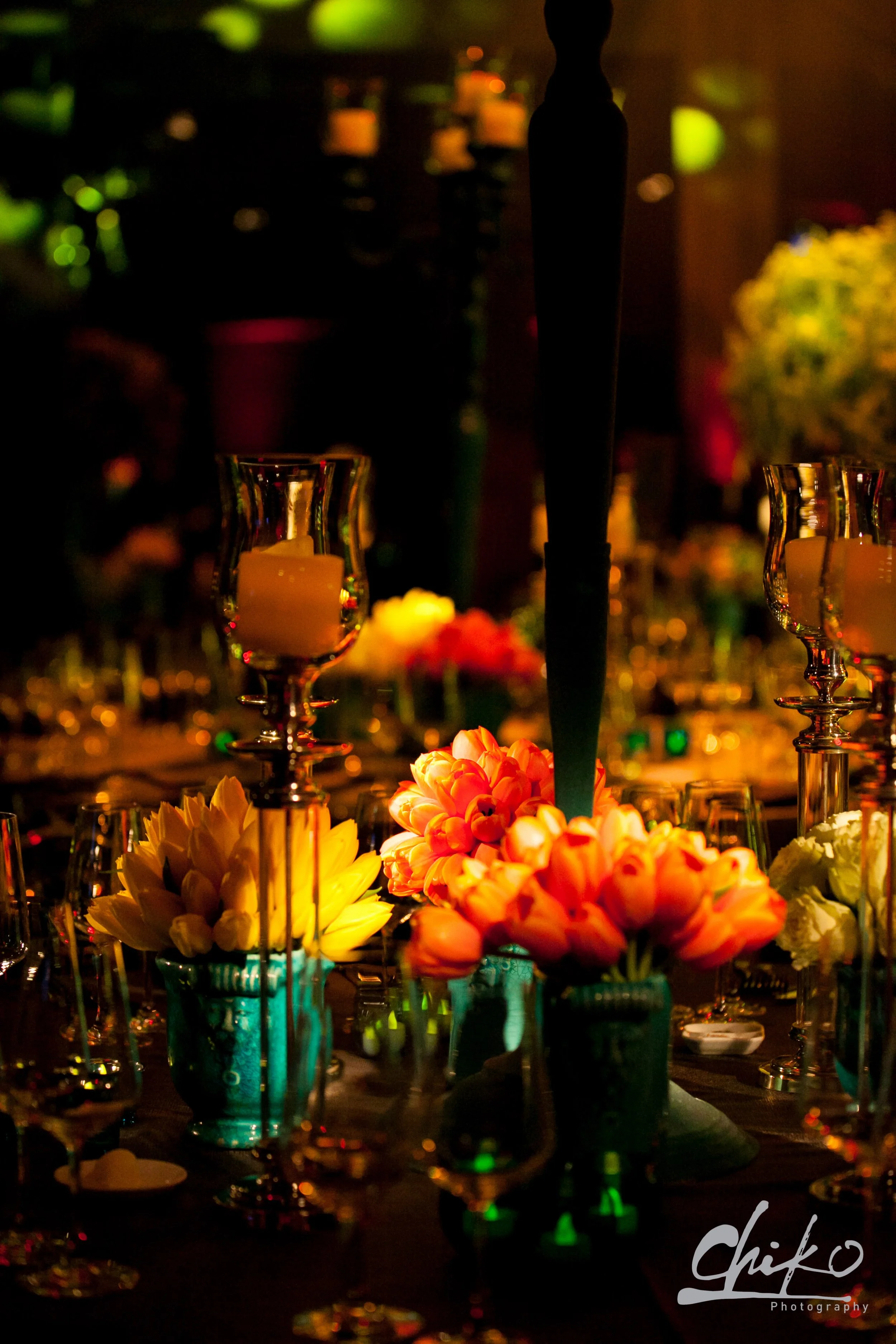 A dimly lit table setting with colorful flowers in tall vases, candles, and wine glasses, creating an elegant ambiance for an event or celebration.