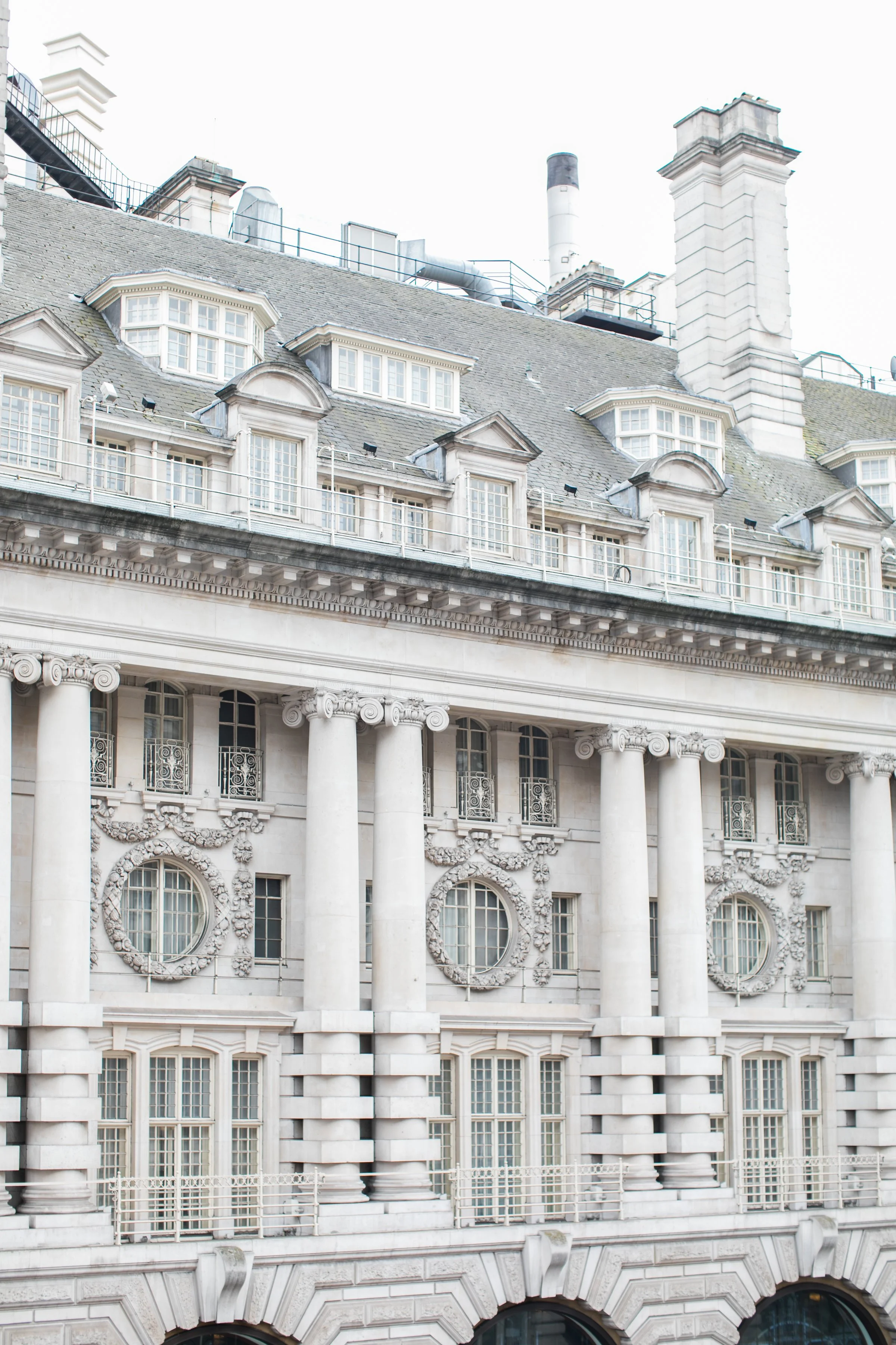 The image shows the upper facade of a historic, ornate building with classical architectural details, including large columns, decorative wreaths around circular windows, and a mansard roof with dormer windows.