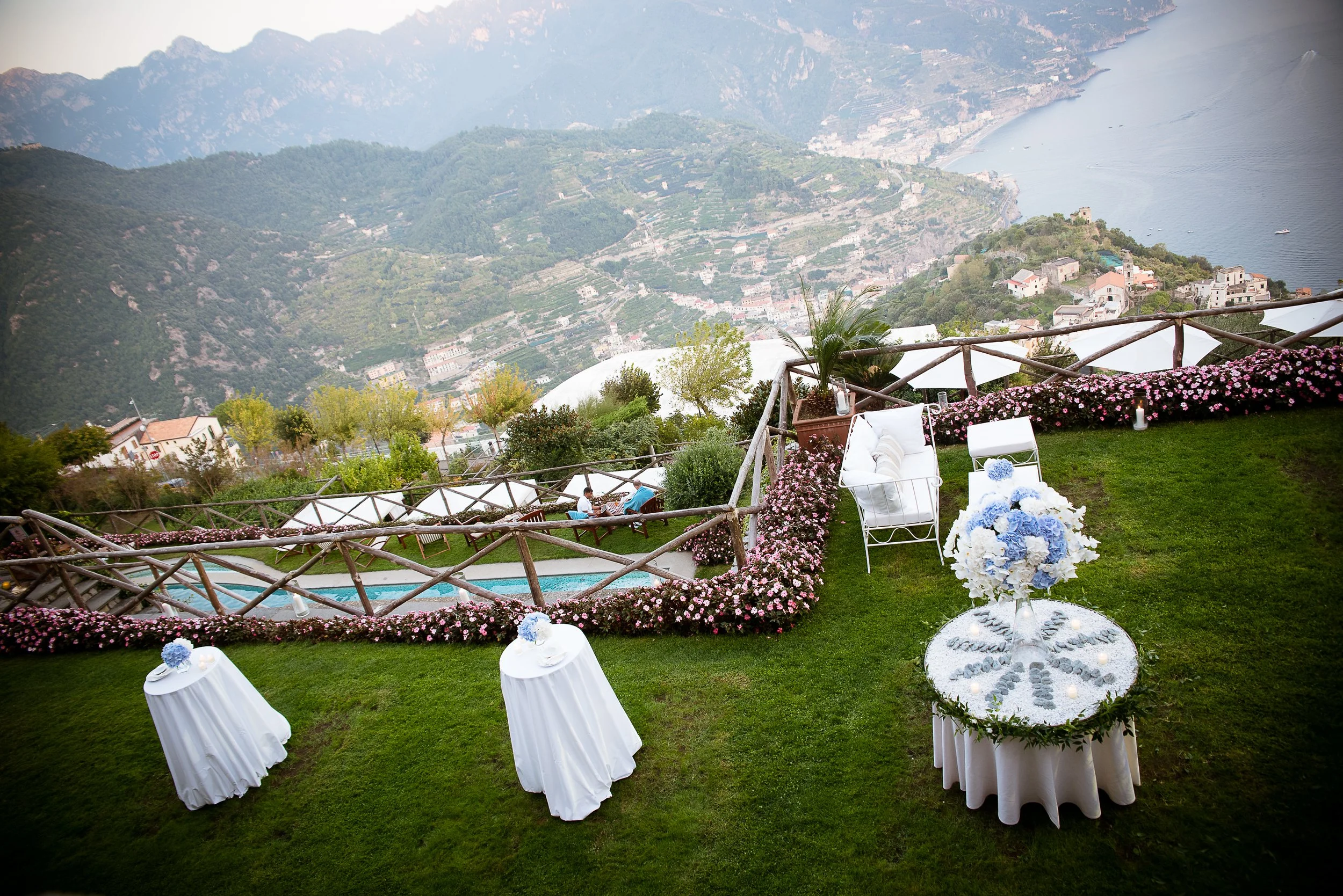 A scenic outdoor event setup on a grassy hillside overlooking a valley with houses and a body of water. The area features white chairs with draped covers and floral decorations, a bouquet of blue and white flowers, and lanterns, with a wooden railing