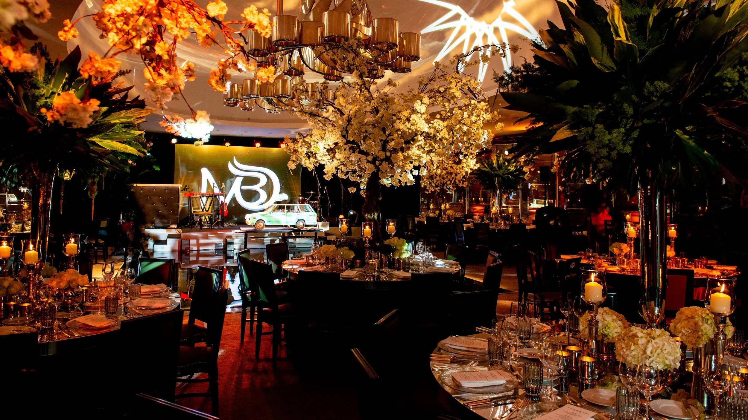 An elegant indoor event space decorated with hanging chandeliers, floral arrangements, and candlelit tables, featuring a stage with a neon 'B' sign, musical instruments, and large floral trees.