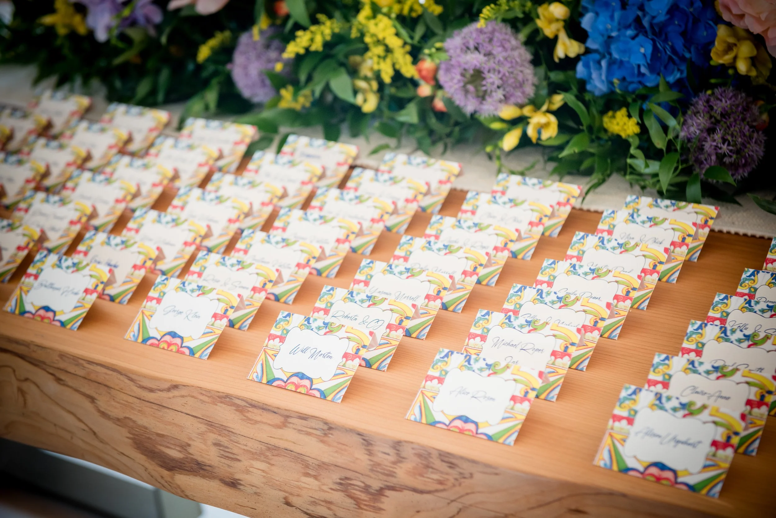 Wedding or event place cards arranged on a wooden table with colorful floral decorations in the background.
